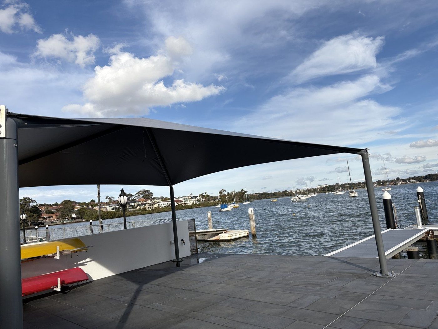 Charcoal canopy on steel posts shading a paved waterfront terrace beside moored boats., Abbotsford