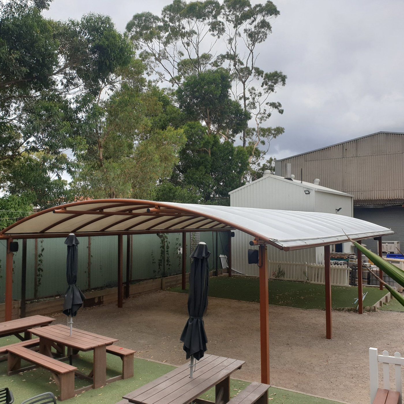Curved cream roof on rust-brown posts shading picnic tables and closed umbrellas on turf., Armidale