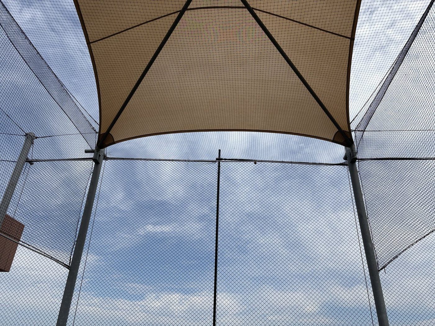 Sand-coloured shade sails tensioned above a tall chain-link fence, seen from below against a cloudy sky., Auburn