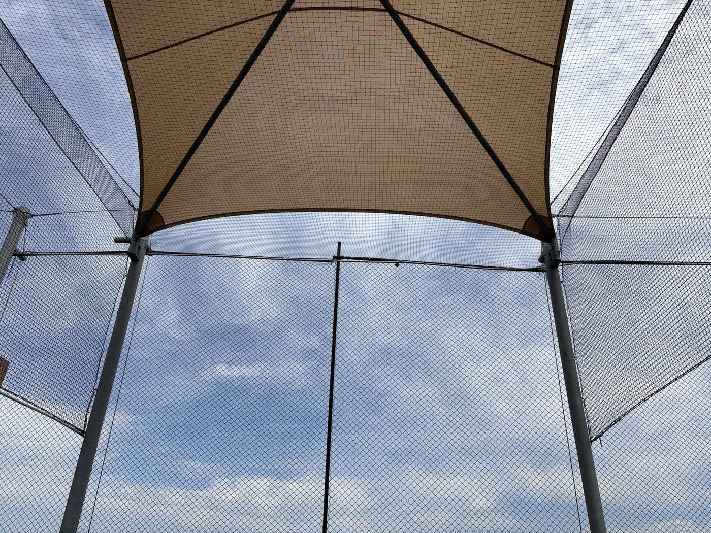 A sand shade sail seen from below inside tall mesh ball netting against a cloudy sky., Auburn