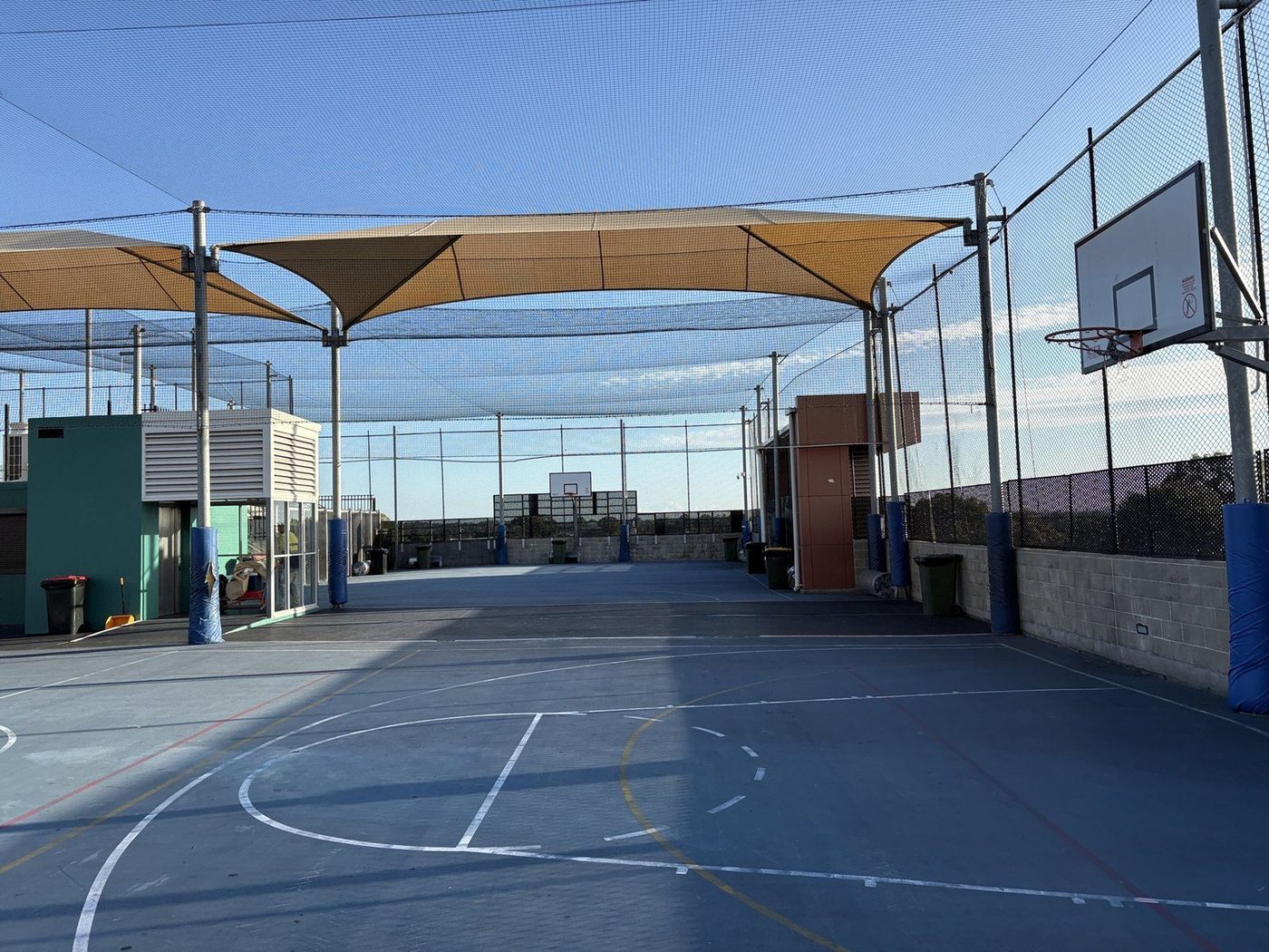 Sand shade sails stretched above a blue rooftop basketball court behind tall ball netting., Auburn