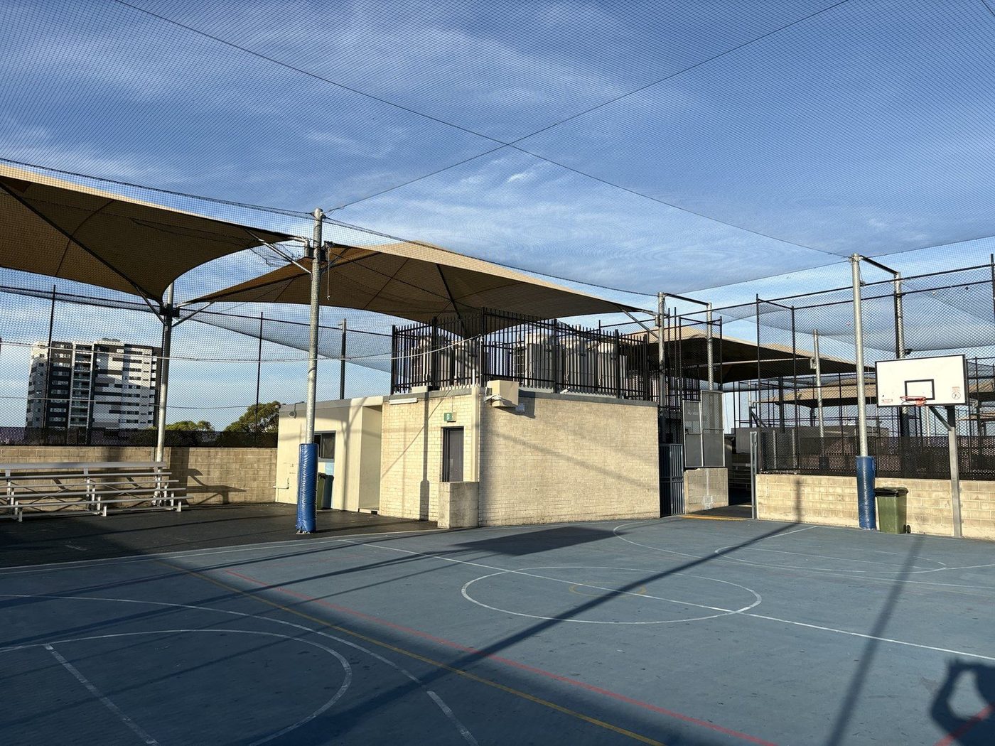 Sand shade sails over a blue outdoor sports court inside tall ball netting, with bleachers to one side., Auburn