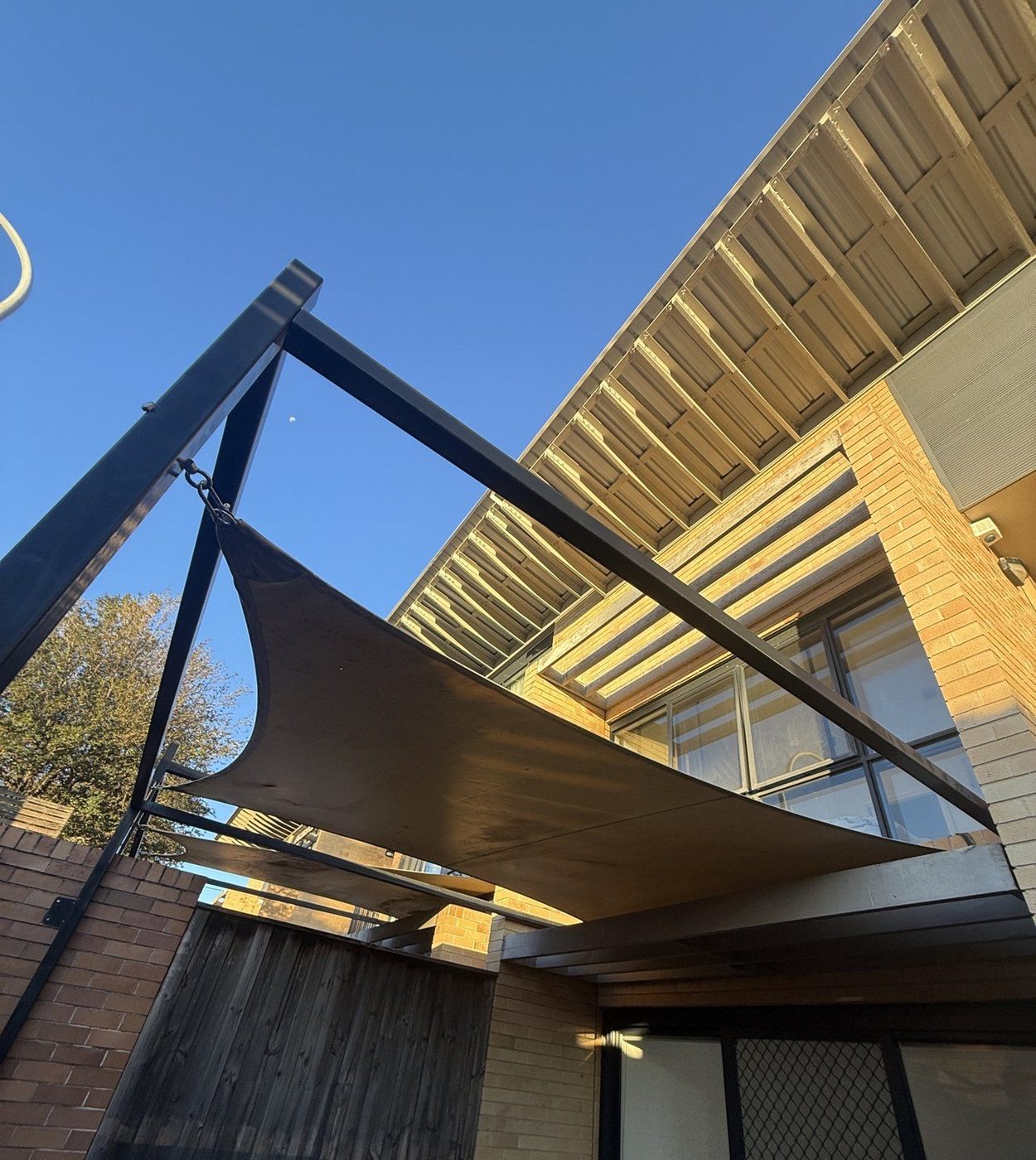 A mocha shade sail on a black steel cantilever frame beside a two-storey brick building in warm light., Carlingford
