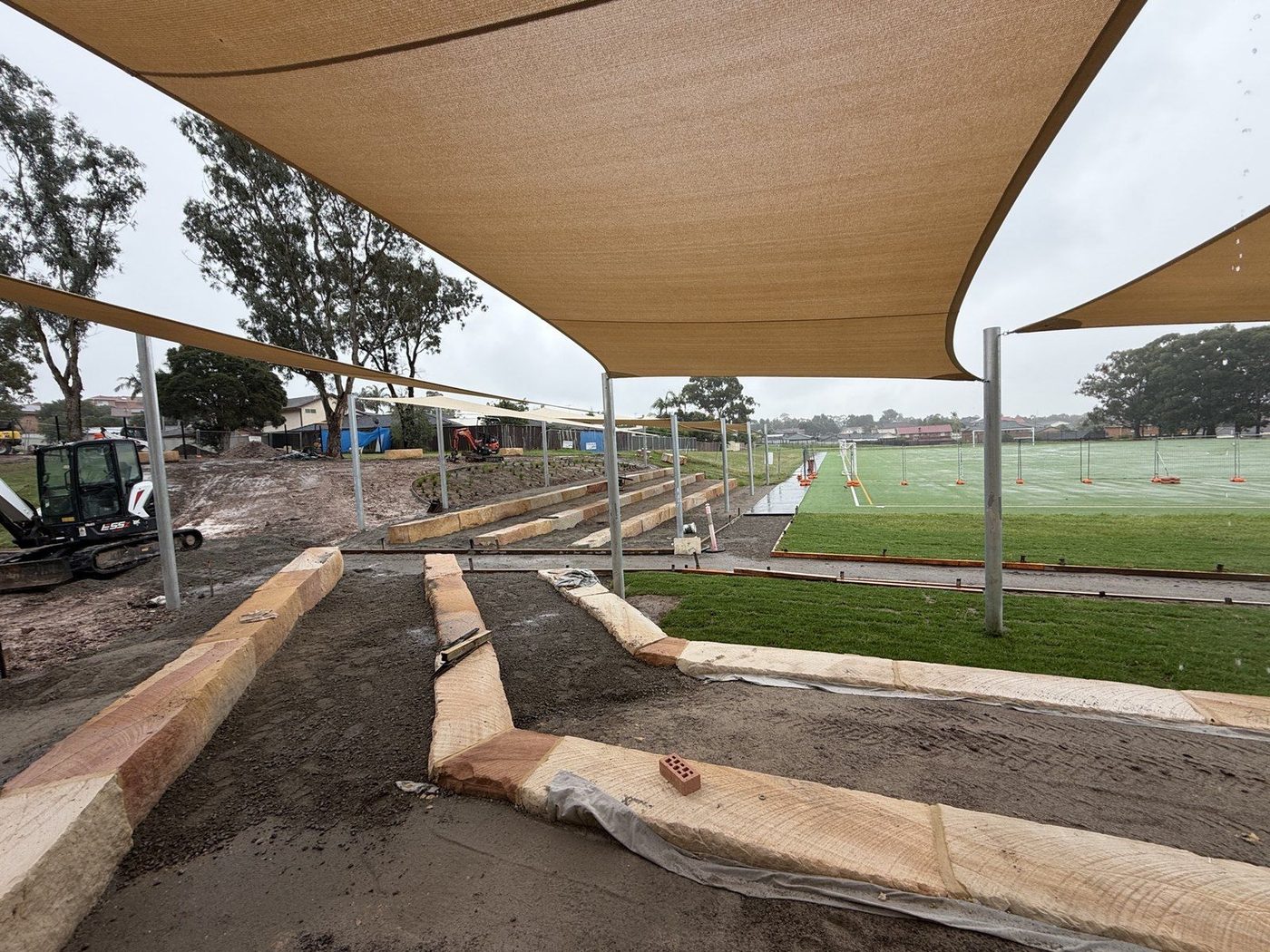 Sand-coloured shade sails over tiered timber seating beside a wet sports field., Casula