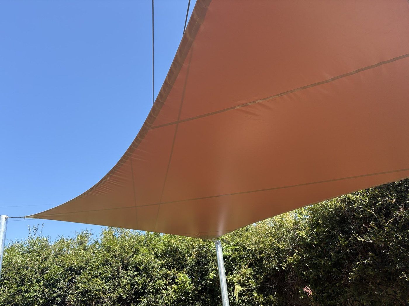 Terracotta sail curving against a clear blue sky above a green hedge., Dapto