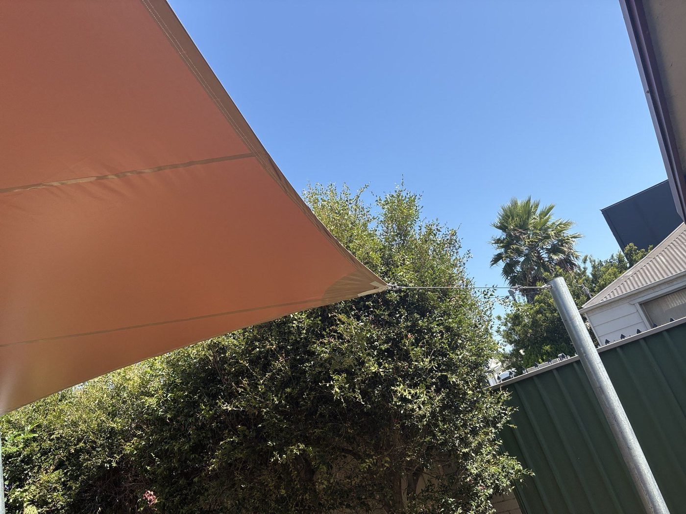 A terracotta shade sail tensioned to a steel post above a back garden under clear blue sky., Dapto