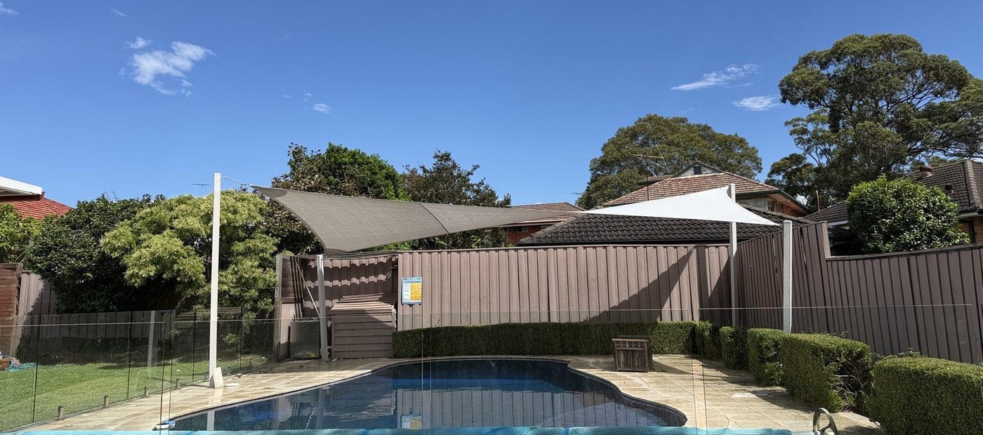 A grey sail and a white sail on steel posts shading the paved edge of a backyard pool., Denistone