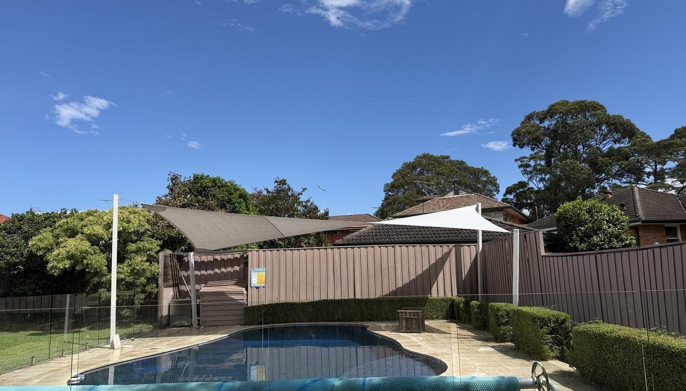 Two shade sails, one taupe and one white, angled on white posts above the paved surround of a backyard pool., Denistone