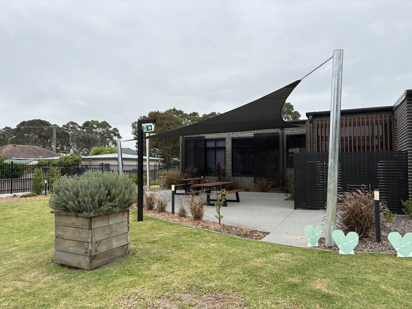 A charcoal shade sail tensioned over a paved courtyard with a picnic table beside a brick building., Doonside