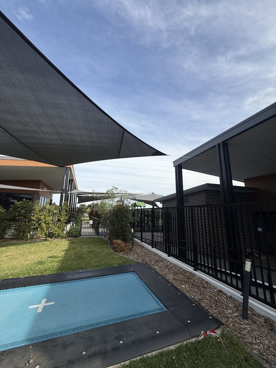 A charcoal shade sail angled over lawn beside an in-ground trampoline in a fenced childcare yard., Doonside
