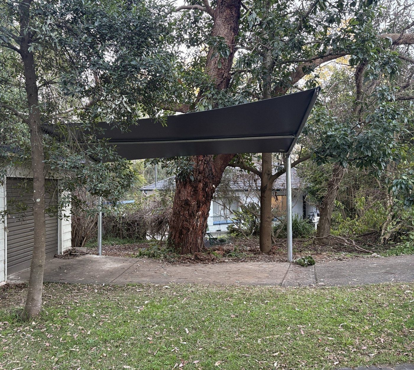 A charcoal shade sail on steel posts over a concrete driveway beside a garage, framed by tall trees., Eastwood