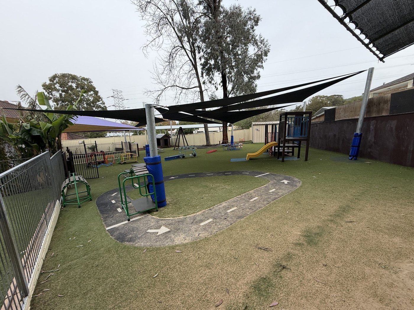 Black shade sails over a childcare play area with a painted road circuit on green turf., Emu Heights