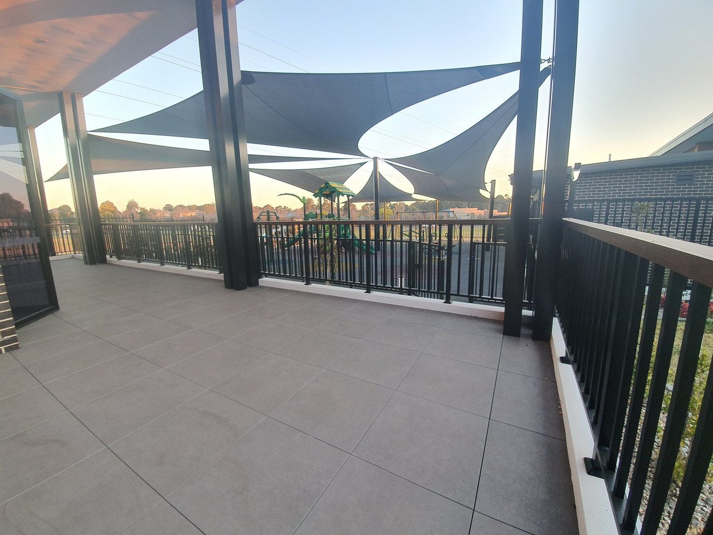 Shade sails in staggered layers above a playground at dusk, seen from a tiled balcony., Emu Heights