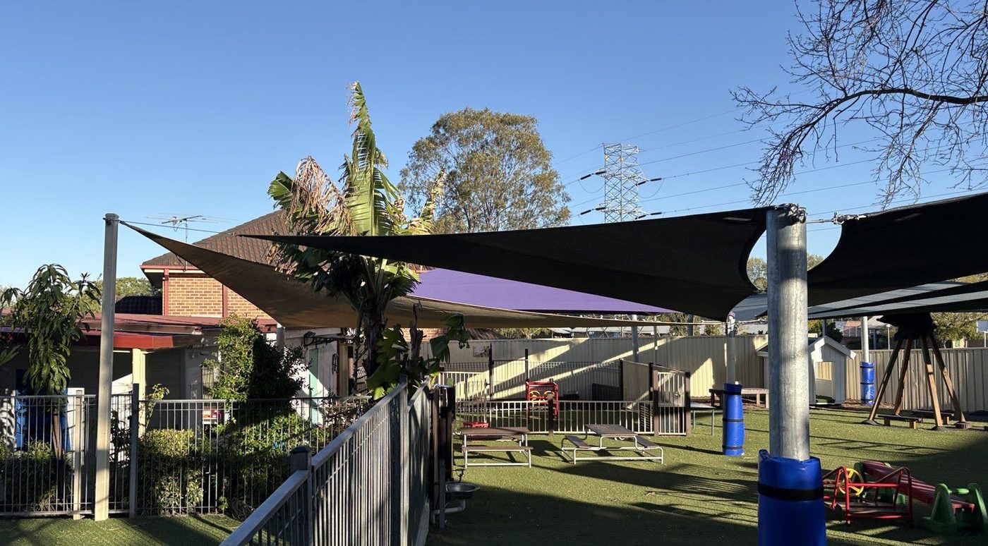 Charcoal and purple shade sails on steel posts above a fenced childcare playground with synthetic turf., Emu Heights