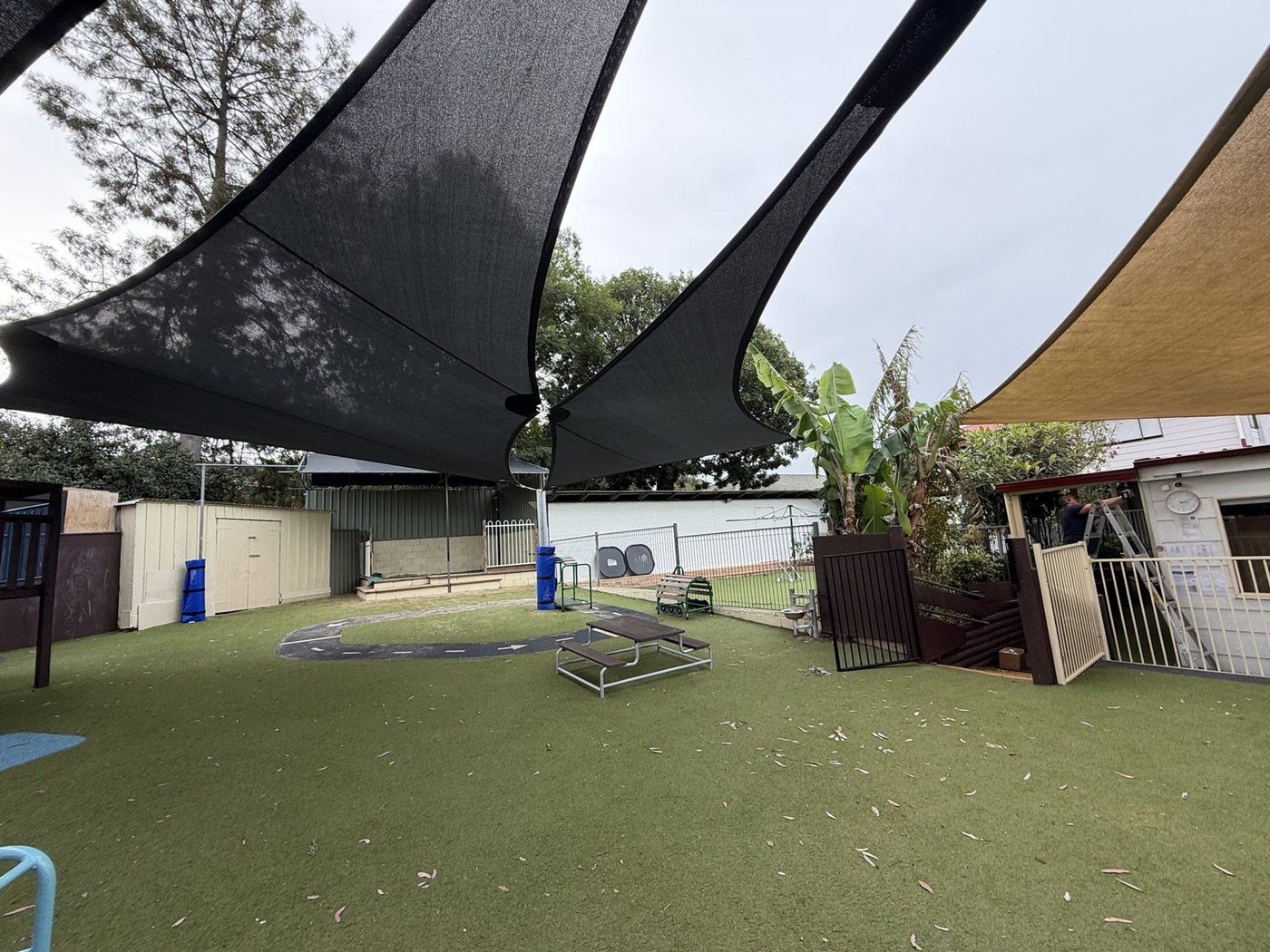 Black shade sails spanning a childcare play yard with synthetic turf and a painted bike track., Emu Heights