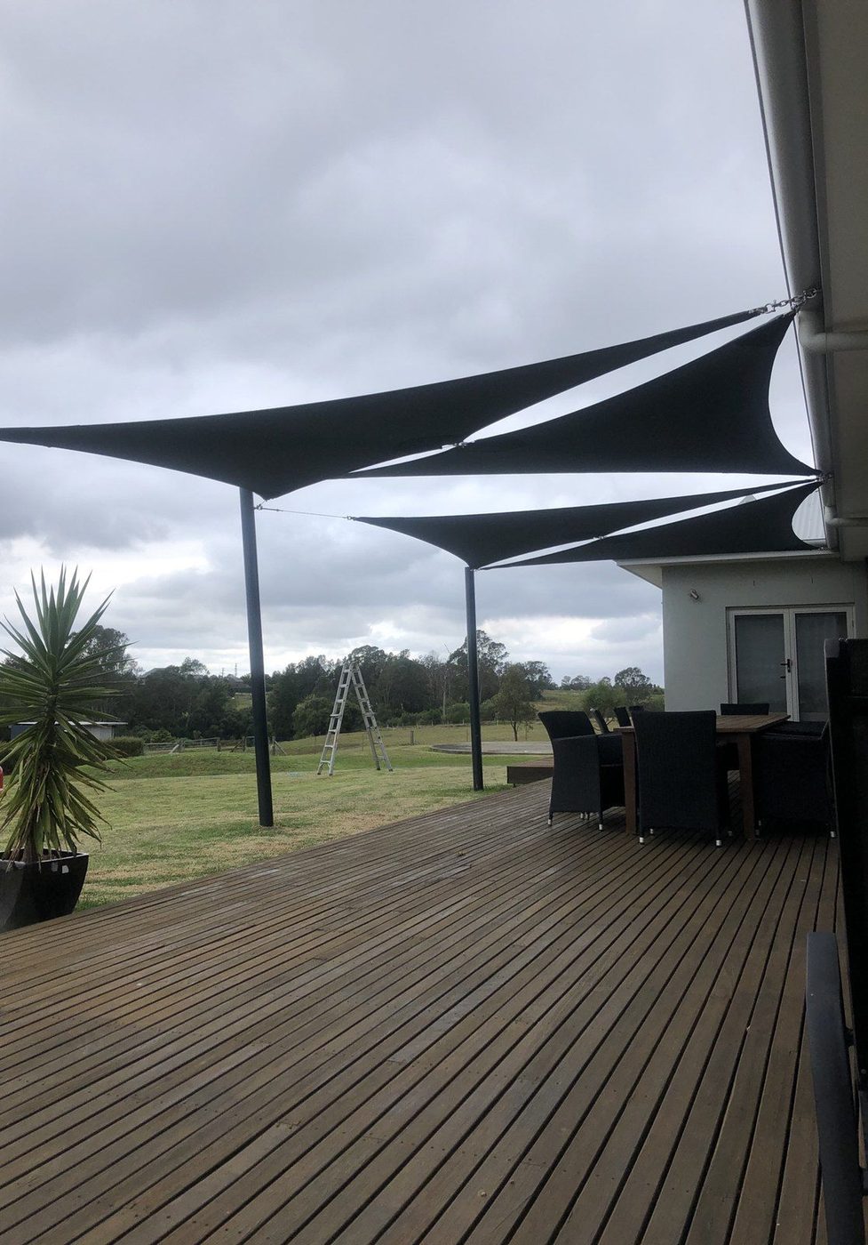 Overlapping charcoal shade sails above a timber deck facing a grassy paddock under a grey sky., Emu Heights