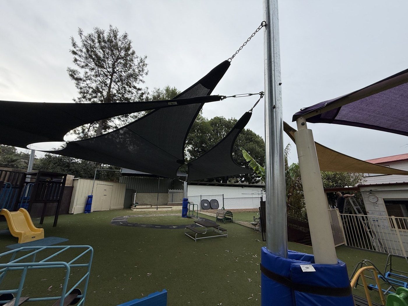 Black shade sails tensioned by chain to a galvanised post over a childcare playground., Emu Heights