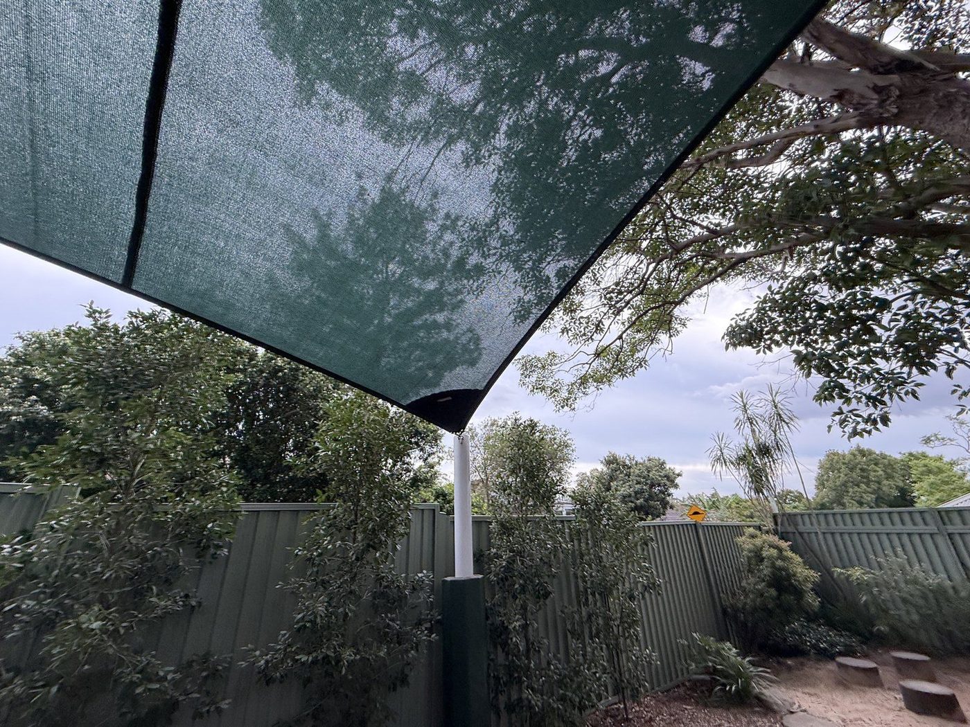 A dark green shade sail pulled tight to a white steel post above a garden and fence line., Ermington
