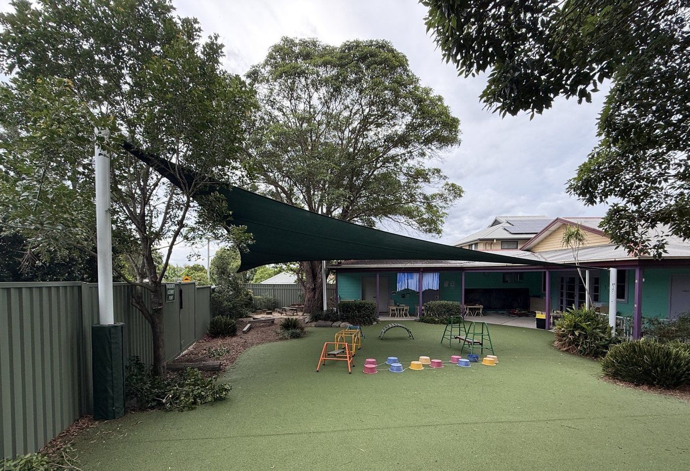 A dark green shade sail angled across a turfed play area on white steel posts under a grey sky., Ermington