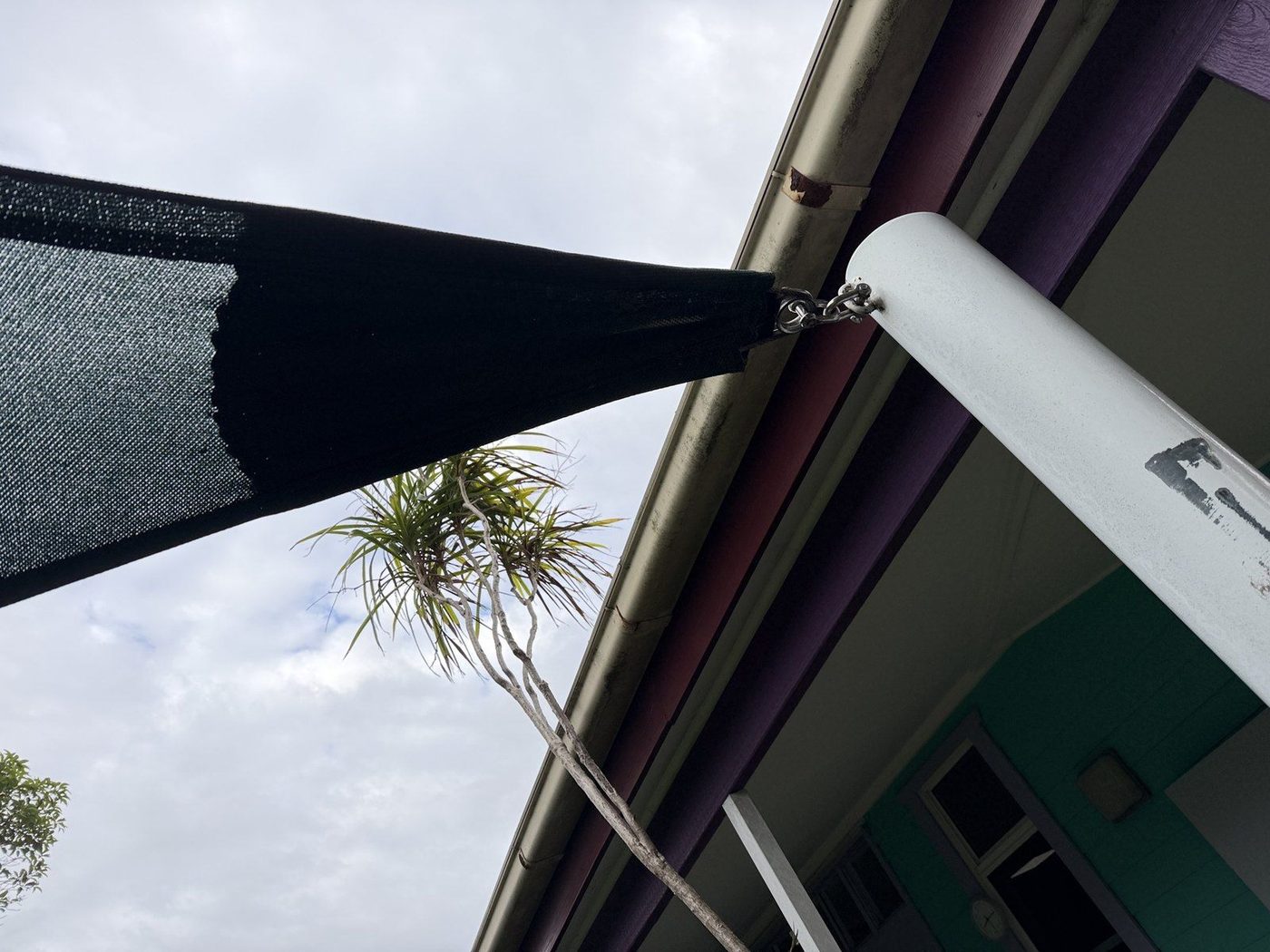 A black shade sail corner chained to the roof edge of a brightly painted building., Ermington
