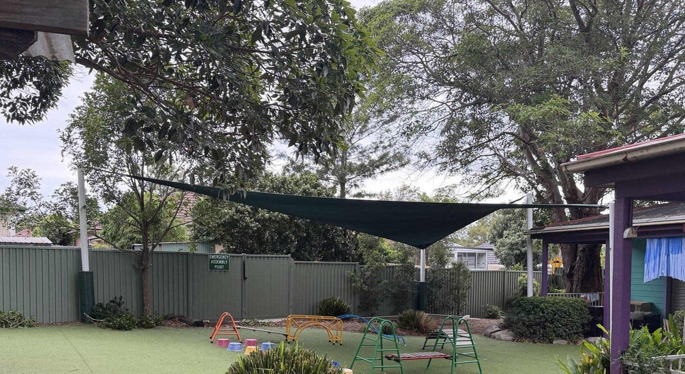 A dark green shade sail stretched above a synthetic grass play yard with small play equipment below., Ermington