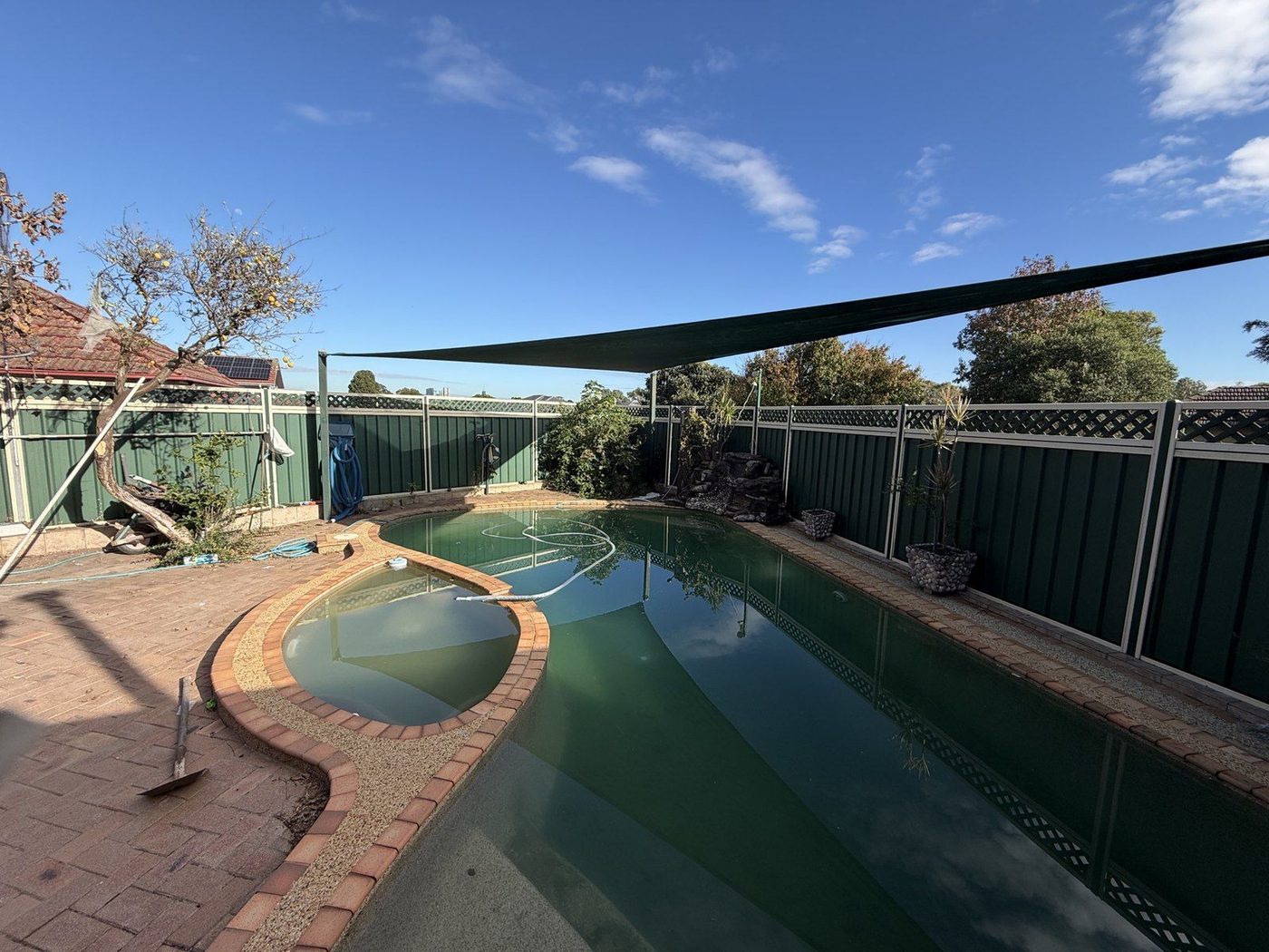 A dark green shade sail stretched above a backyard swimming pool and its paved surround., Ermington