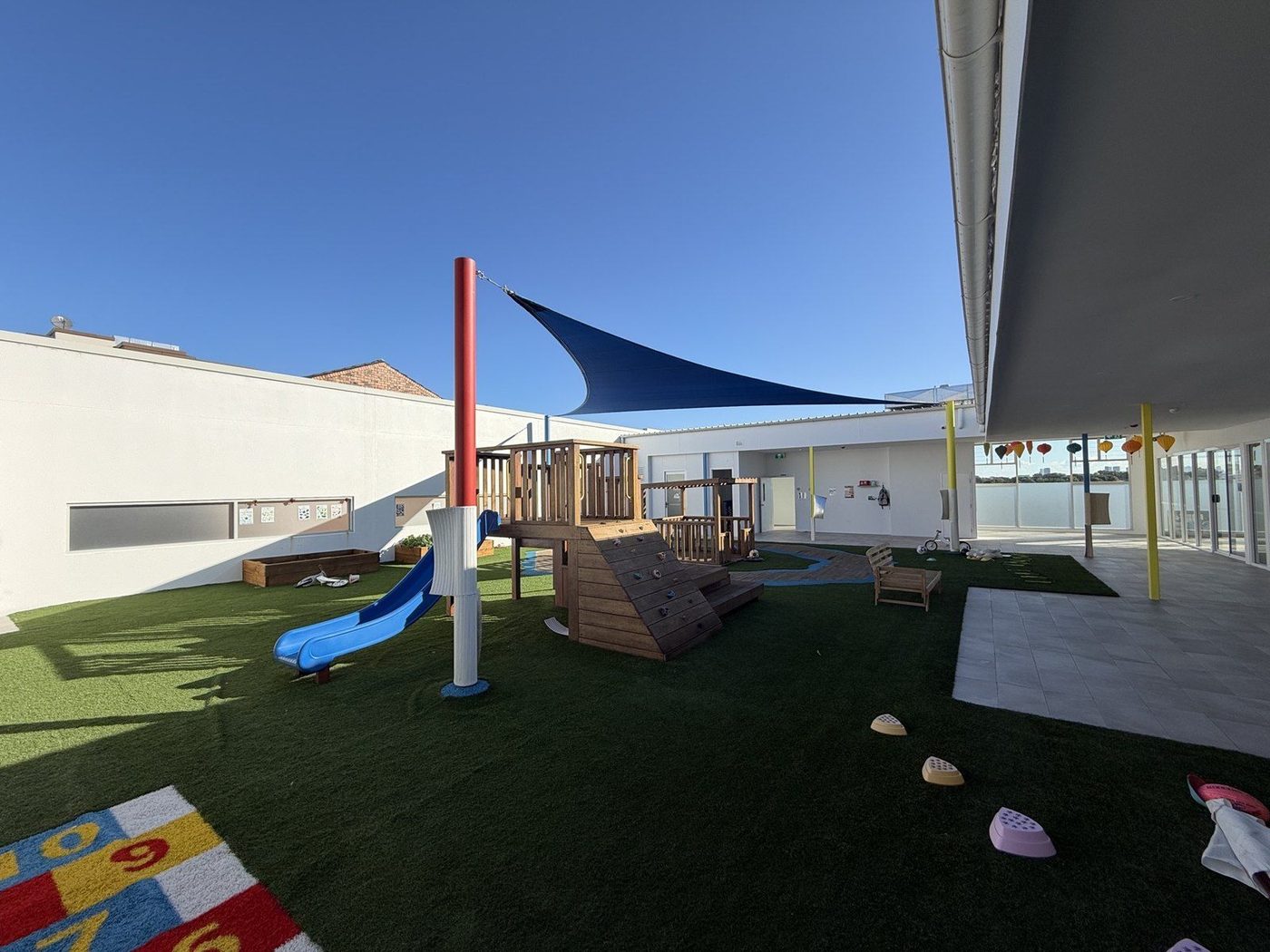 A navy shade sail on a red post above a rooftop childcare play yard with a slide and climbing frame., Five Dock