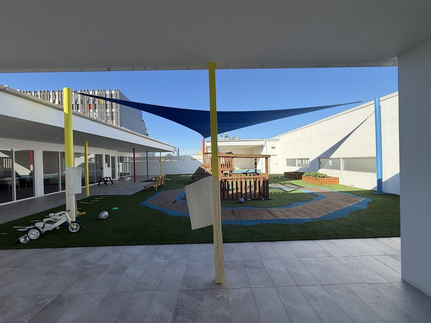 A blue shade sail on yellow posts over a synthetic turf courtyard playground with a timber cubby., Five Dock