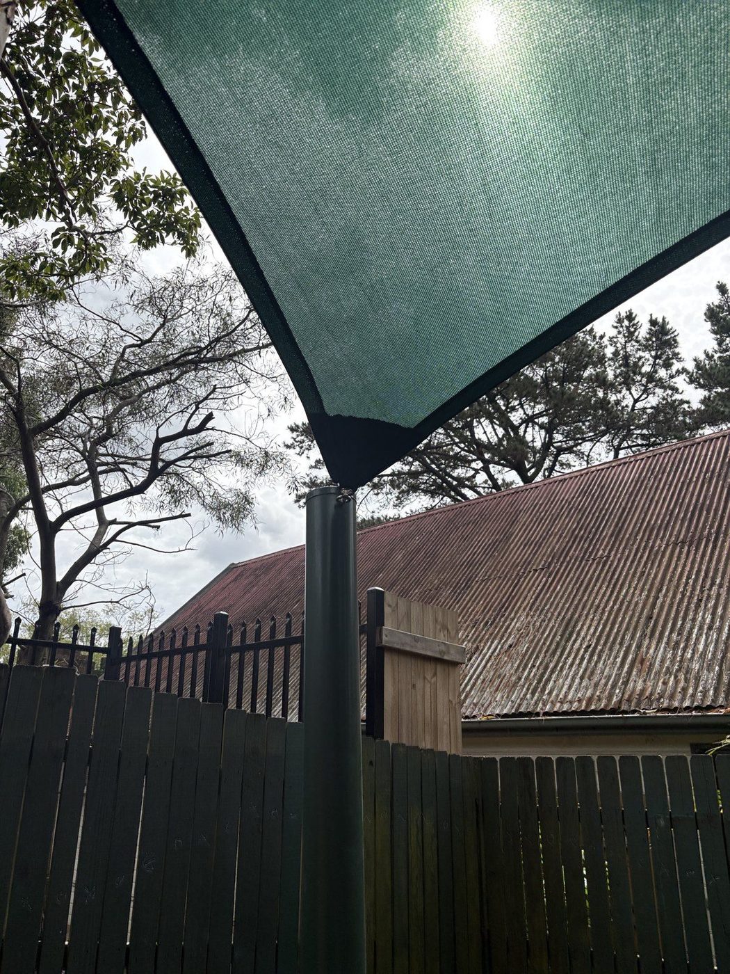 Green shade sail corner tensioned to a dark steel post above a paling fence, with an old tin roof behind., Forestville