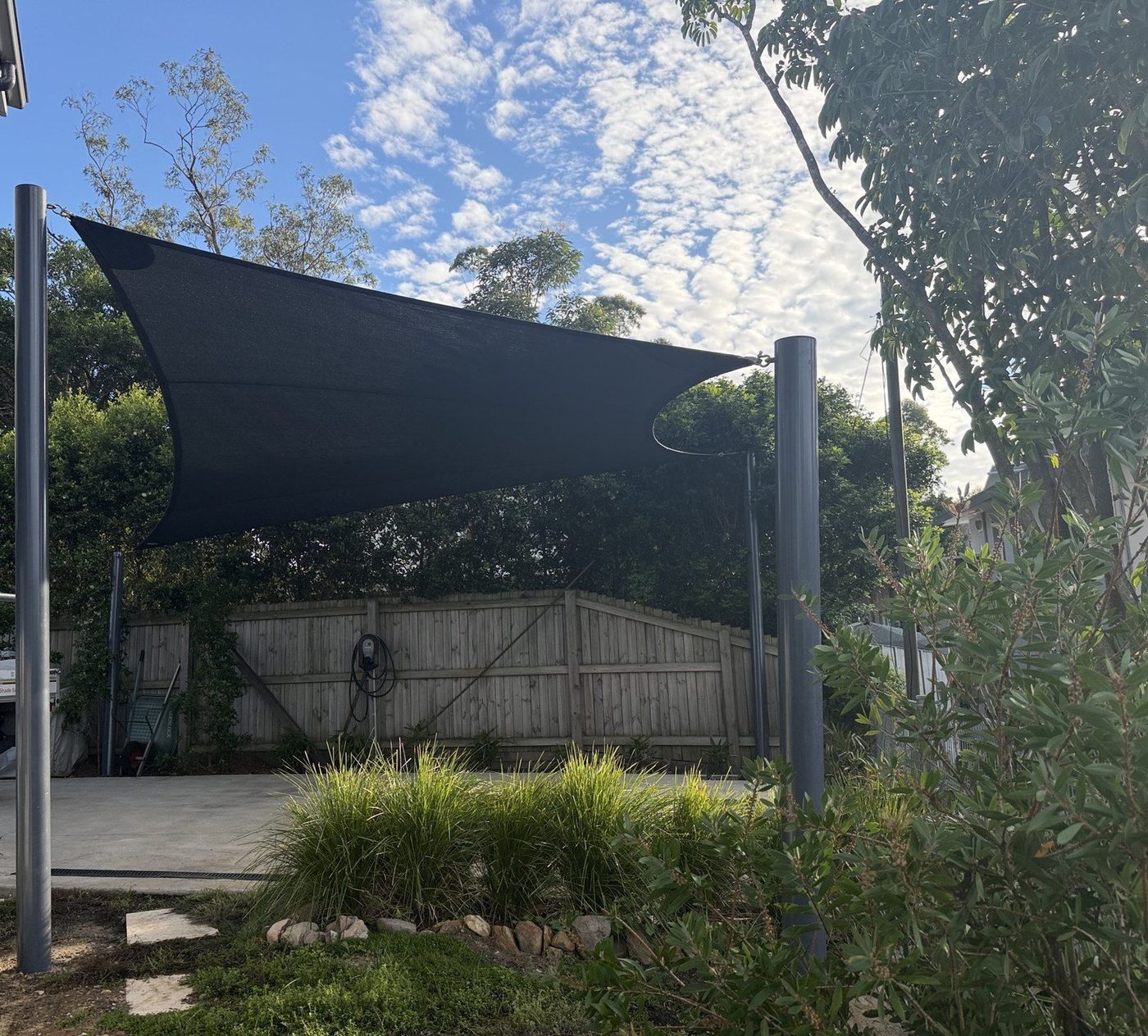 A charcoal square shade sail on tall steel posts over a concrete driveway beside a timber paling fence., Forestville