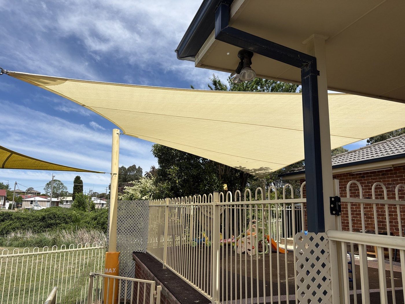 Sand-coloured shade sails stretched from a verandah roof over a fenced play area with play equipment, Goulburn