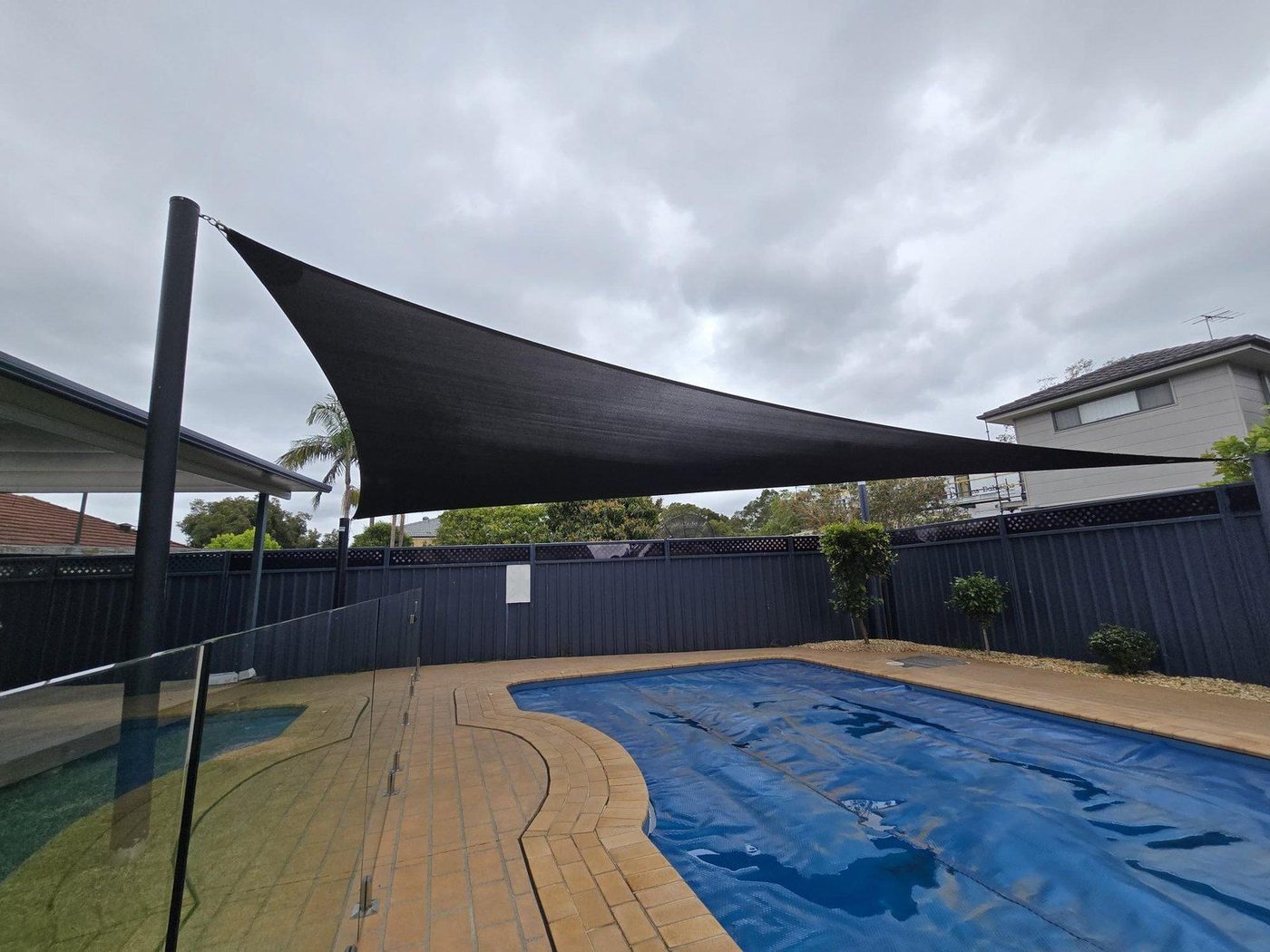 Black shade sail on steel posts stretched above a covered backyard swimming pool under grey cloud., Hammondville