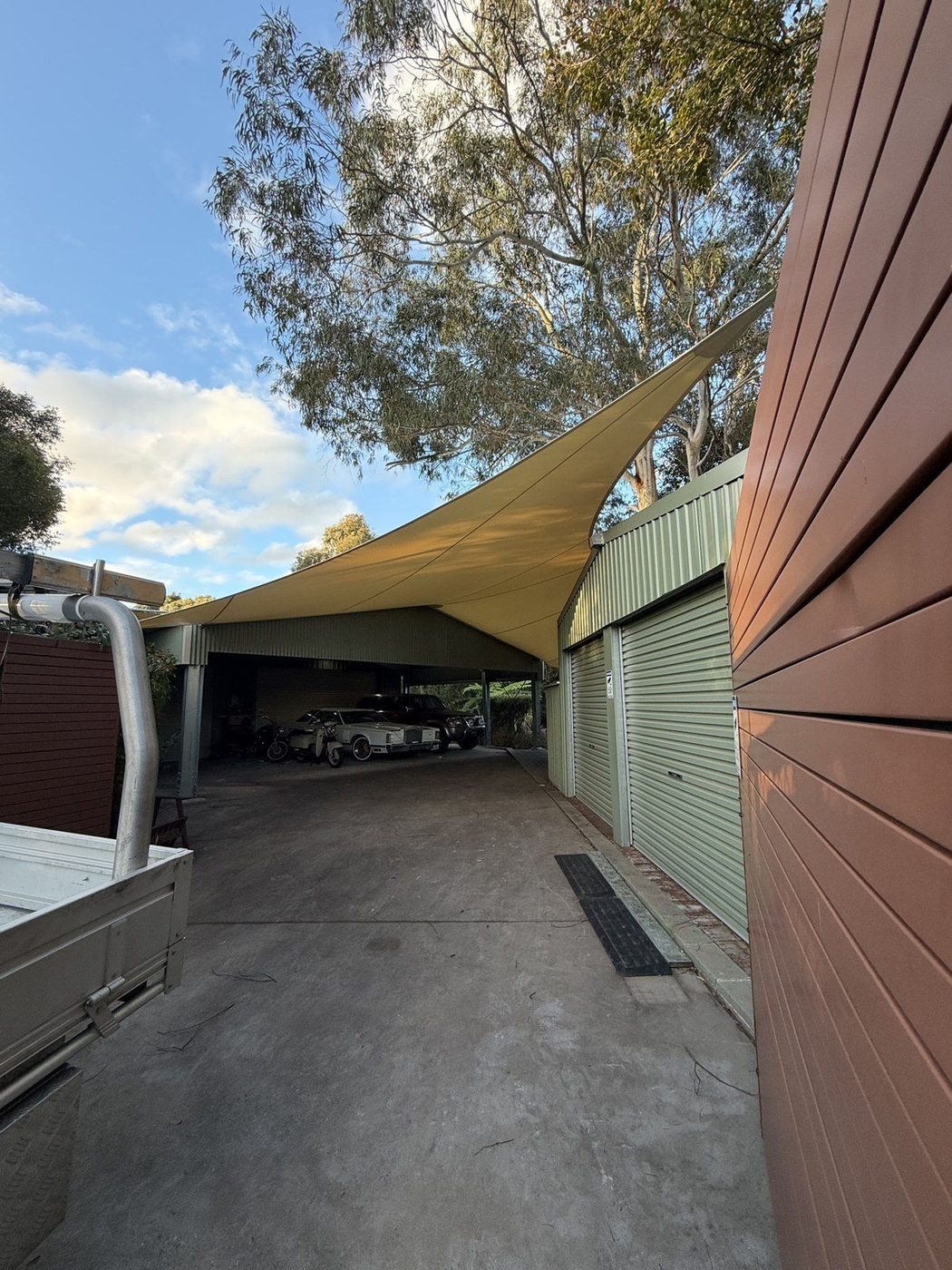 Sand-coloured shade sail stretched over a concrete driveway leading to an open carport at a home., Ingleside