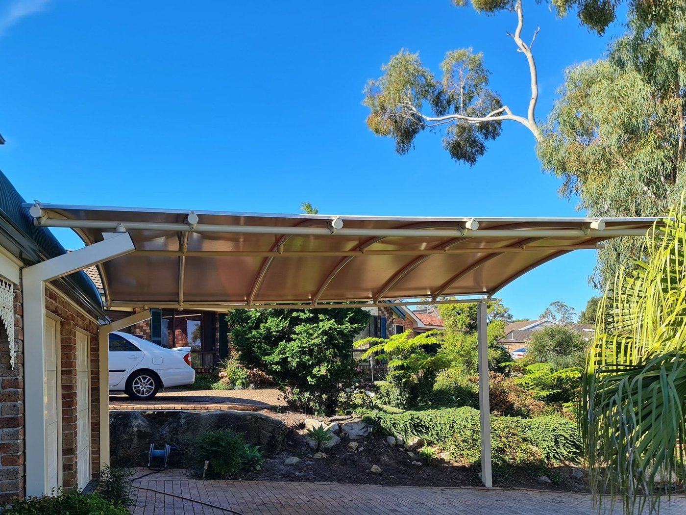 Curved cream carport roof on slim posts over a brick-paved home driveway under clear blue sky., Kellyville