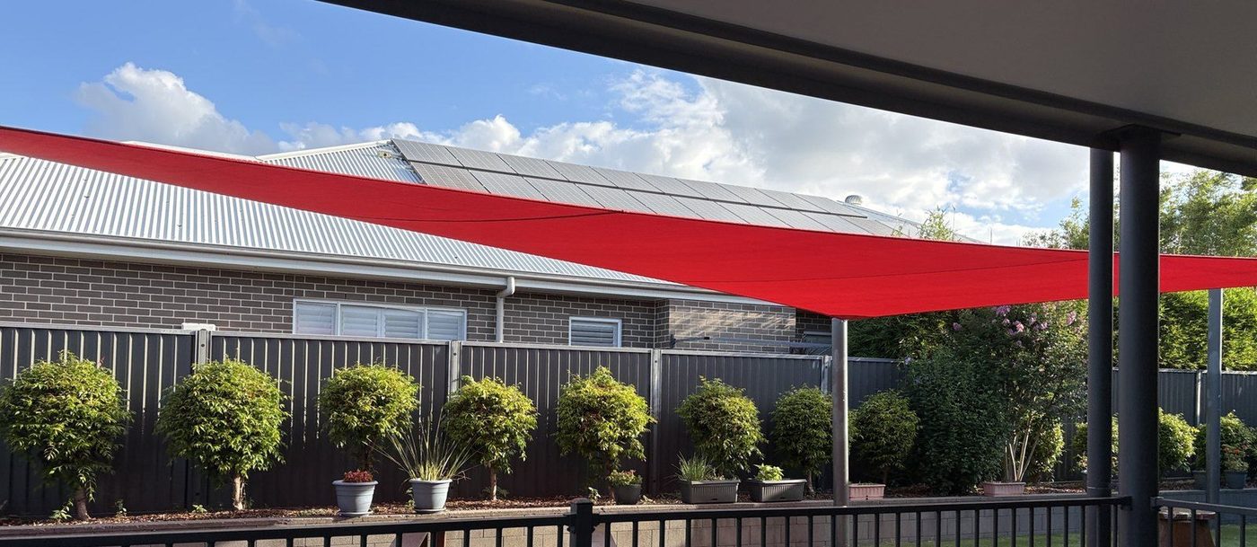 A red rectangular shade sail stretches across a fenced backyard, seen from under a patio., Kellyville