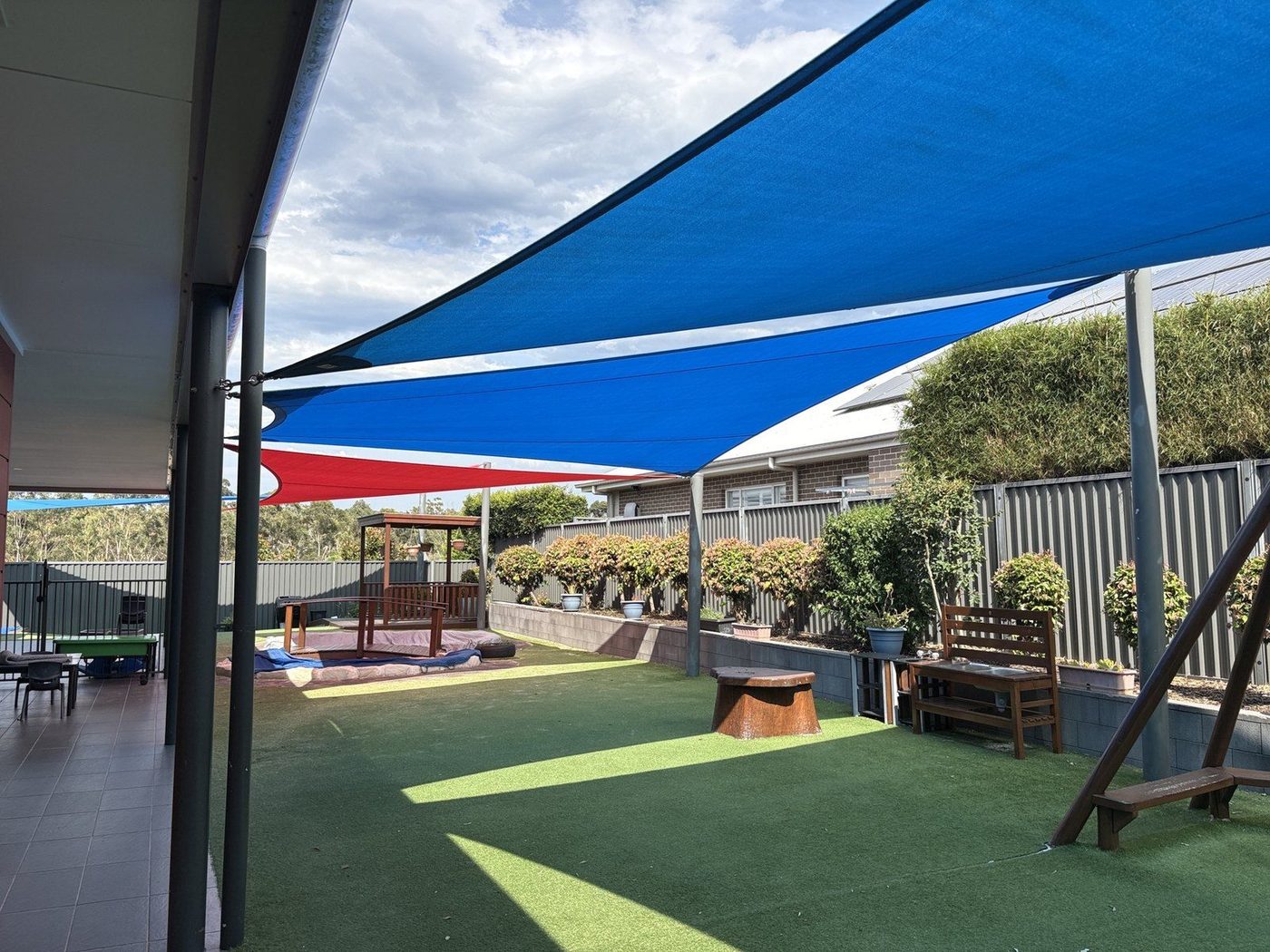 Blue and red shade sails stretched over a synthetic-turf playground with a sandpit and timber play fort., Kellyville