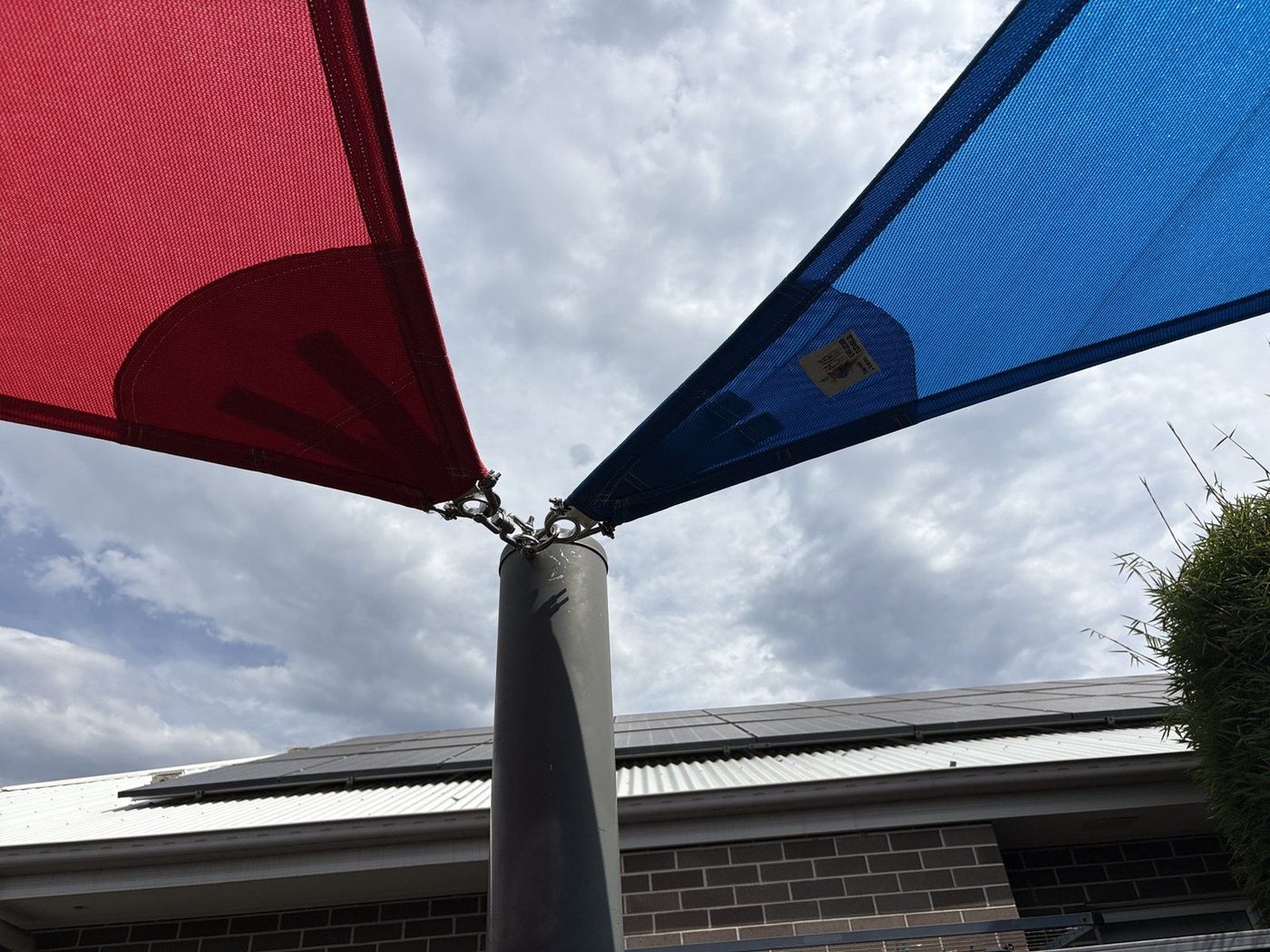 Blue and red shade sails tensioned off a galvanised post head against a cloudy sky., Kellyville