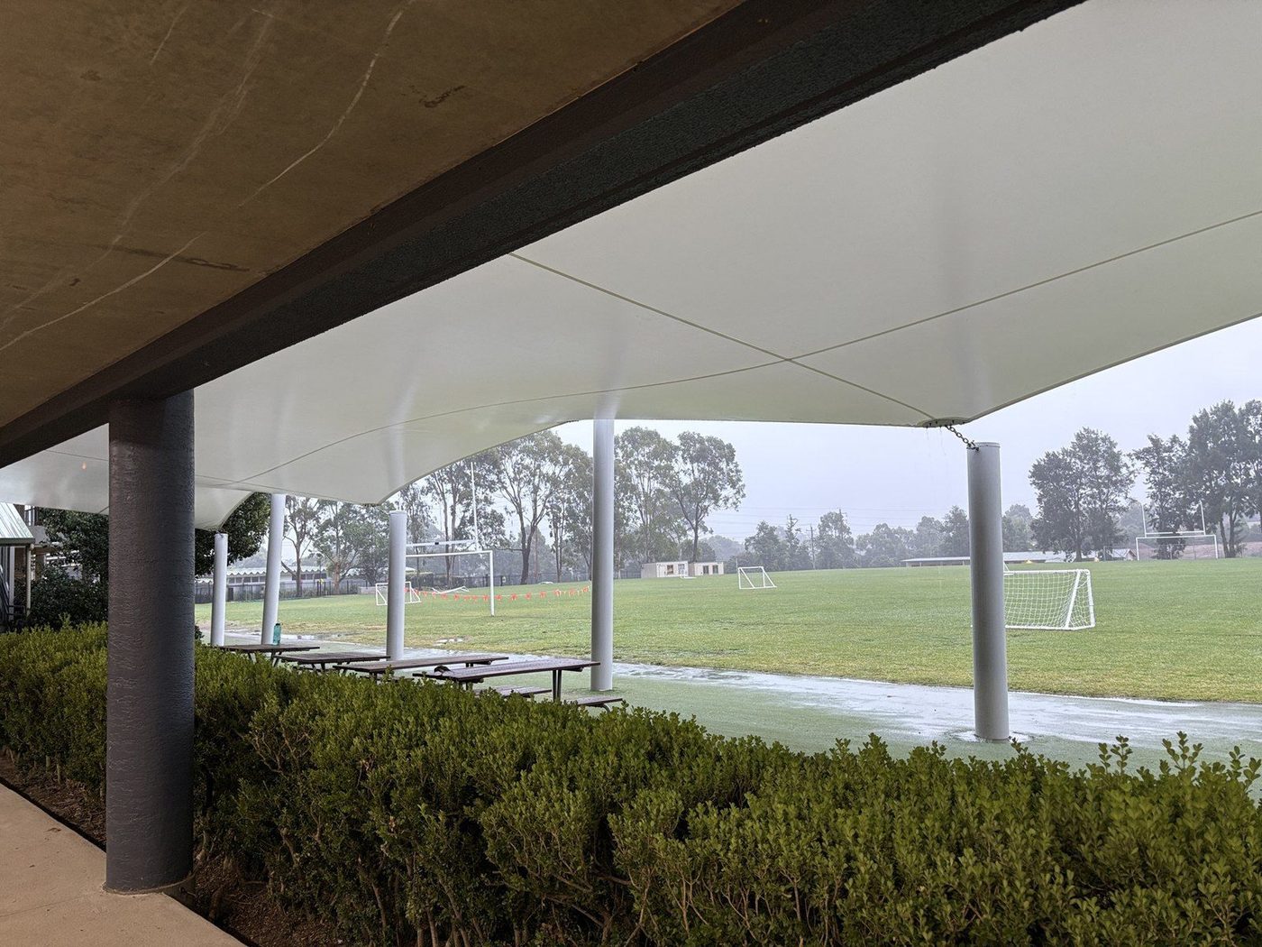 A white membrane canopy on round posts sheltering bench seating beside a wet grass sports field, Kellyville