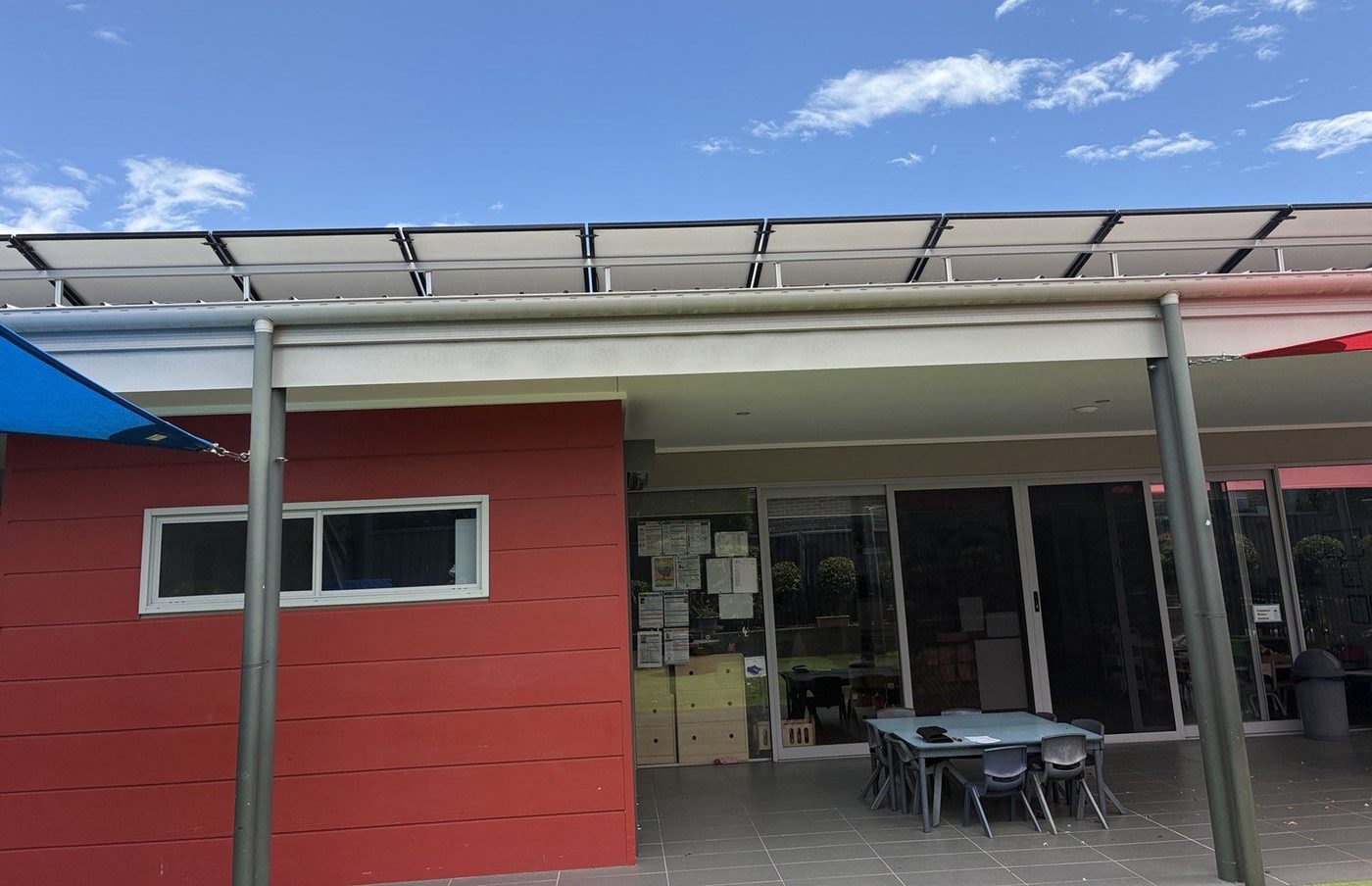 Pale flat-panel roof on slim posts shading a paved childcare courtyard with tables and chairs, Kellyville