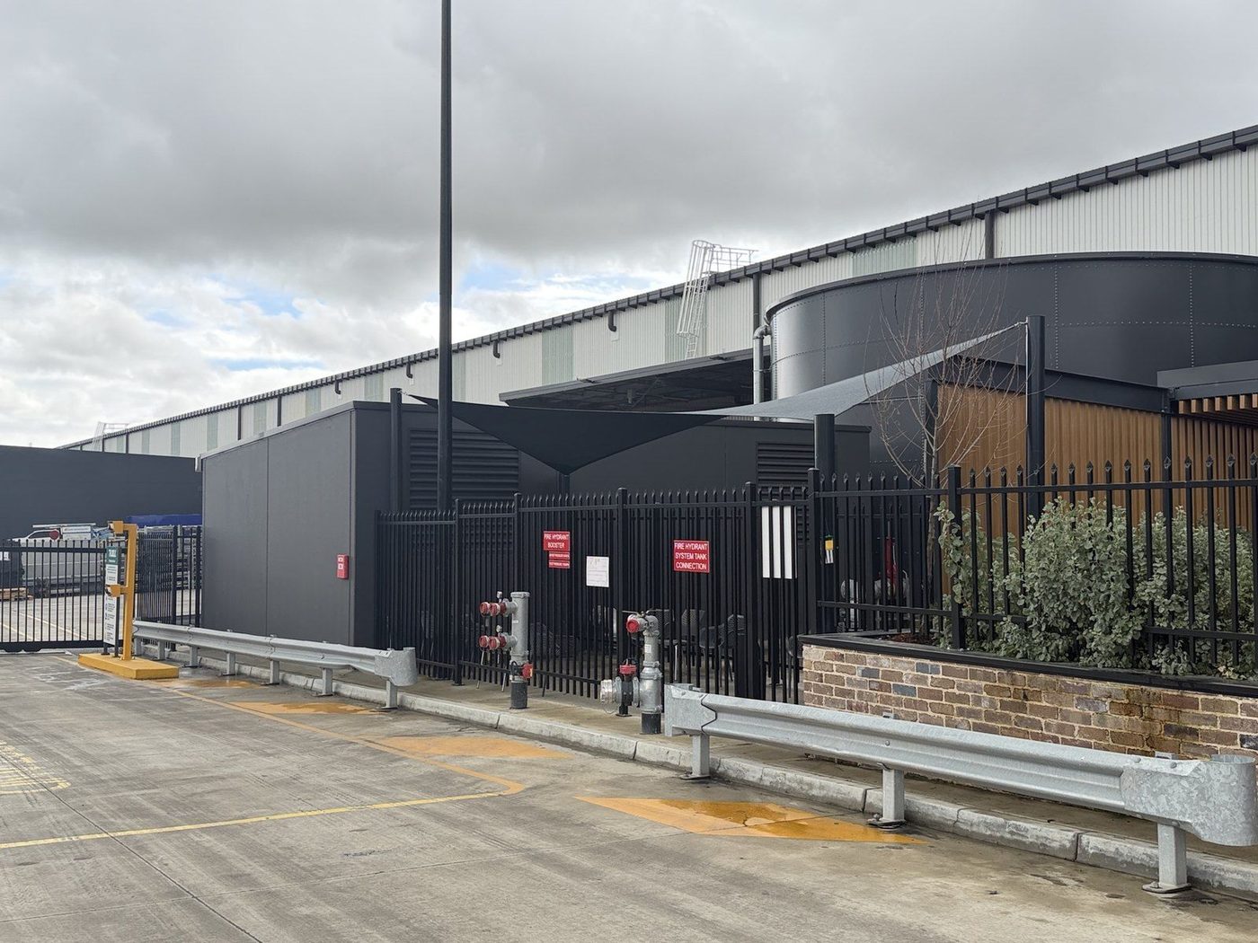 Charcoal shade sail on steel posts above a fenced industrial yard beside a warehouse., Kemps Creek