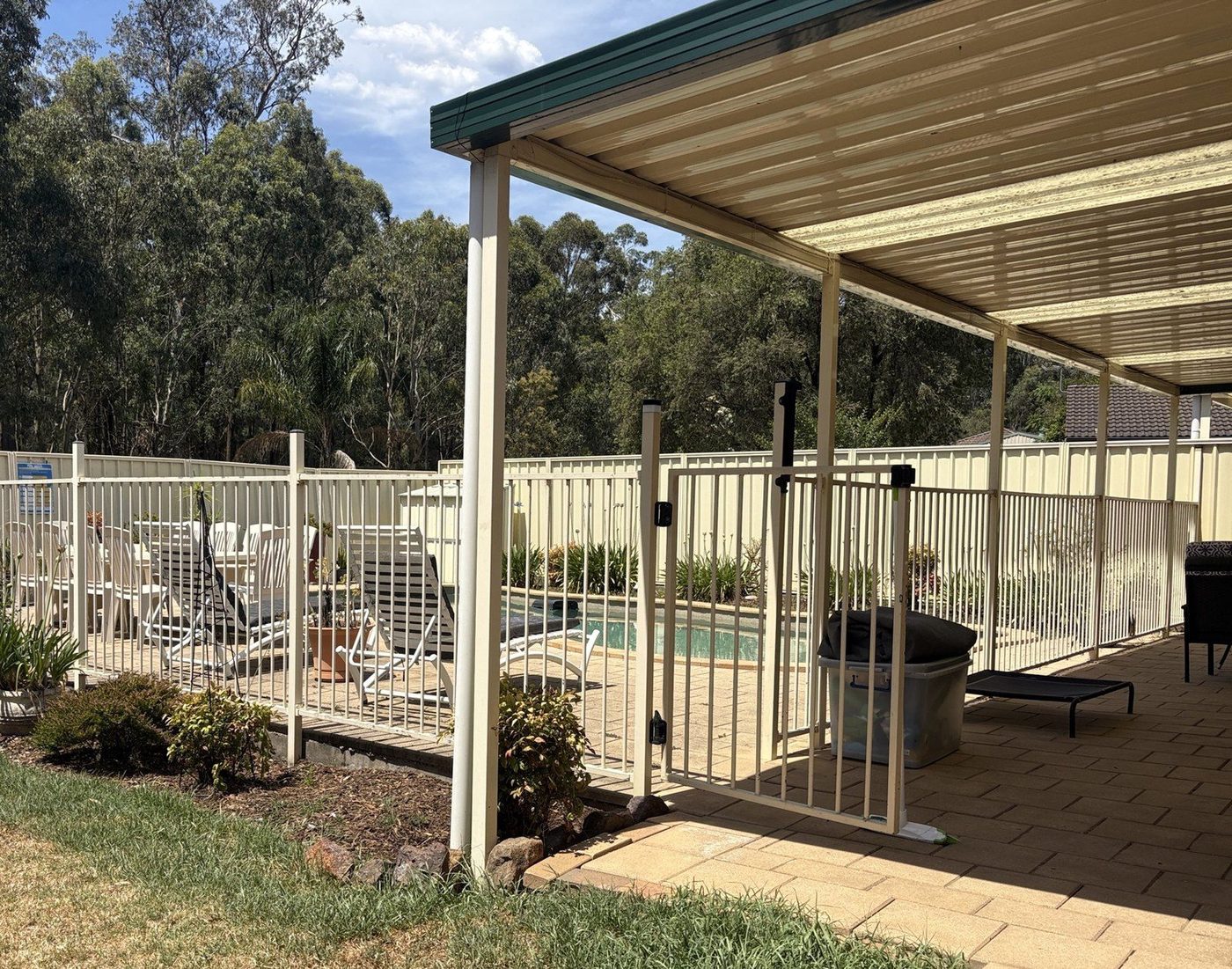 A cream metal patio roof on white posts running along a paved area beside a fenced pool., Kings Park