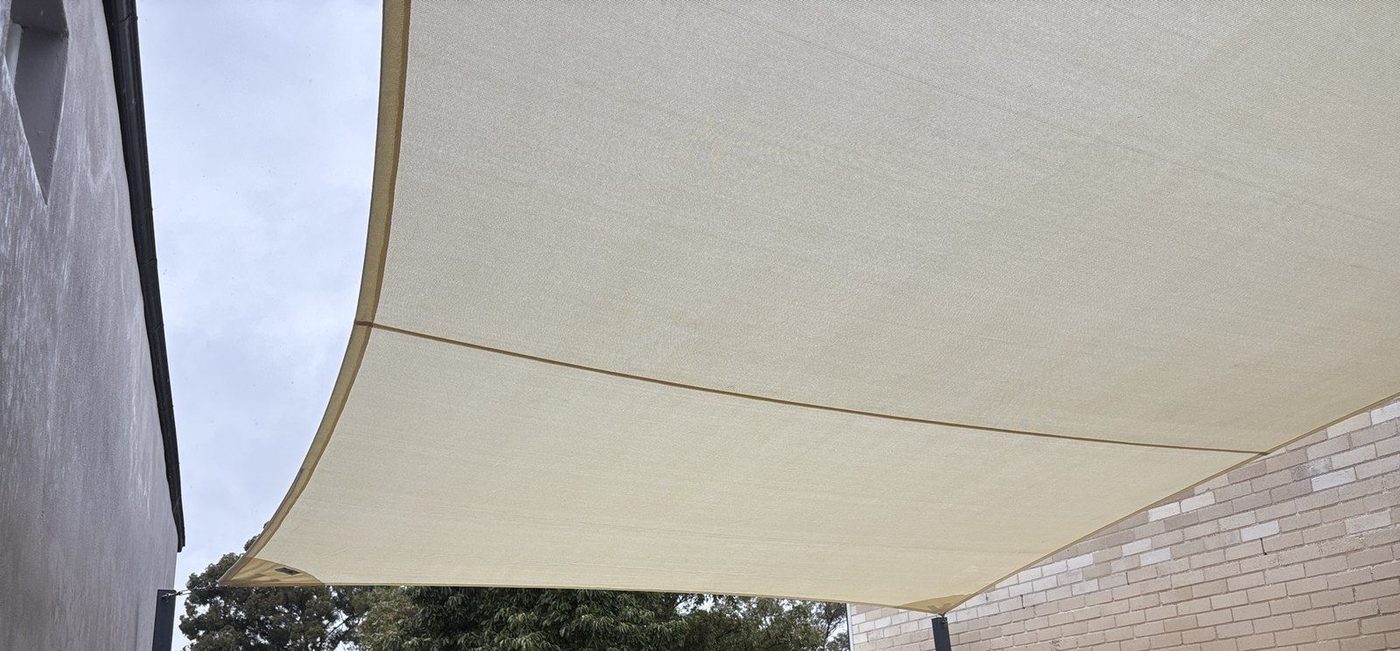A sand shade sail seen from below, spanning between a grey rendered wall and a light brick wall., Leichhardt