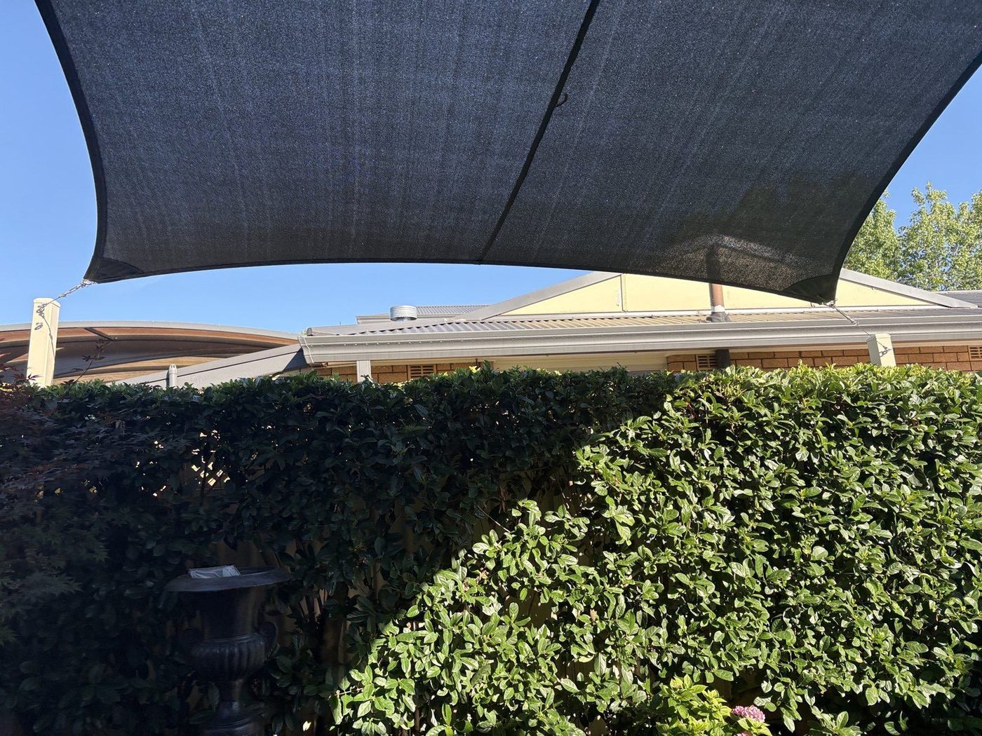 Underside of a charcoal shade sail against clear blue sky above a clipped green garden hedge., Leonay