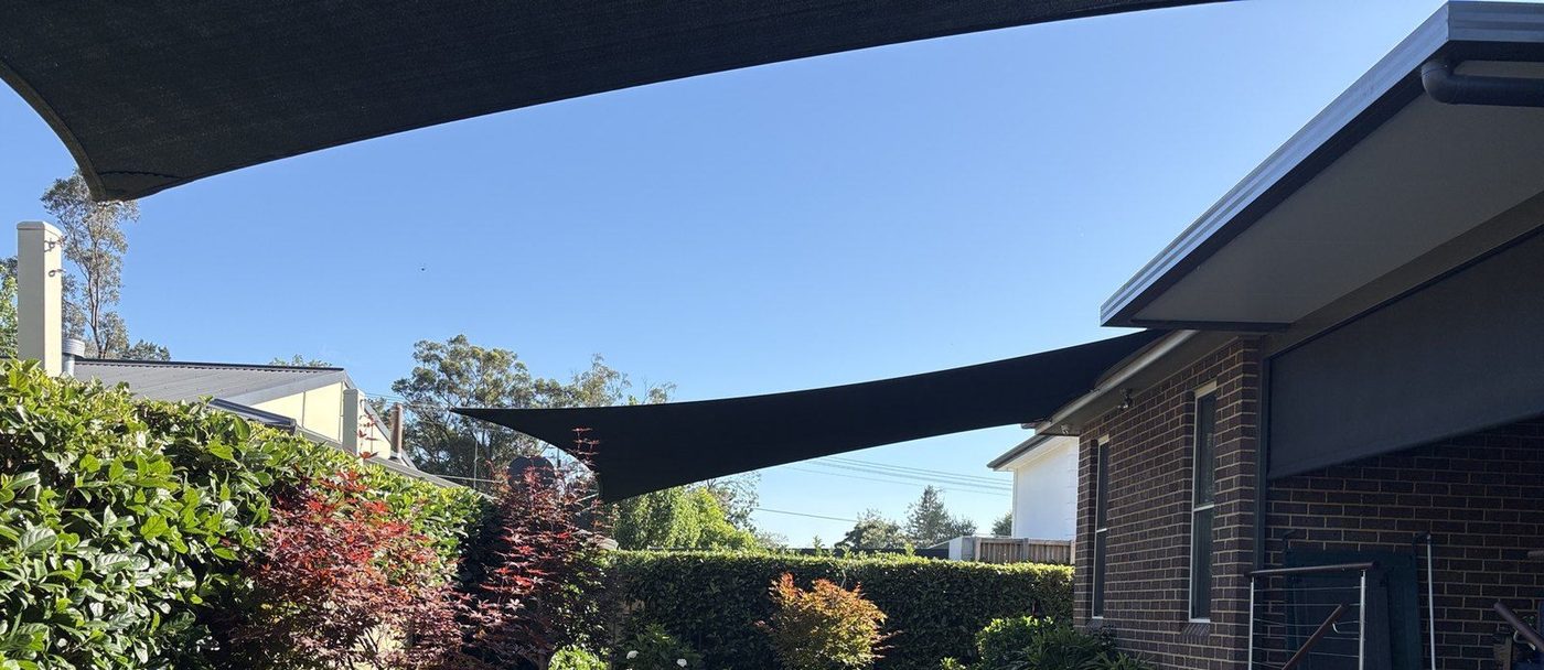 Two charcoal shade sails tensioned above a hedged back garden beside a dark brick home., Leonay