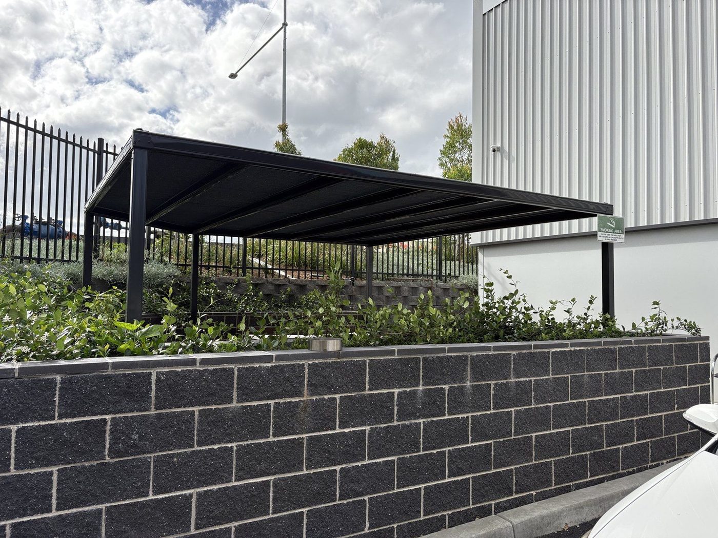 Flat black shade cloth roof on steel posts beside a white commercial building and garden beds., Leppington