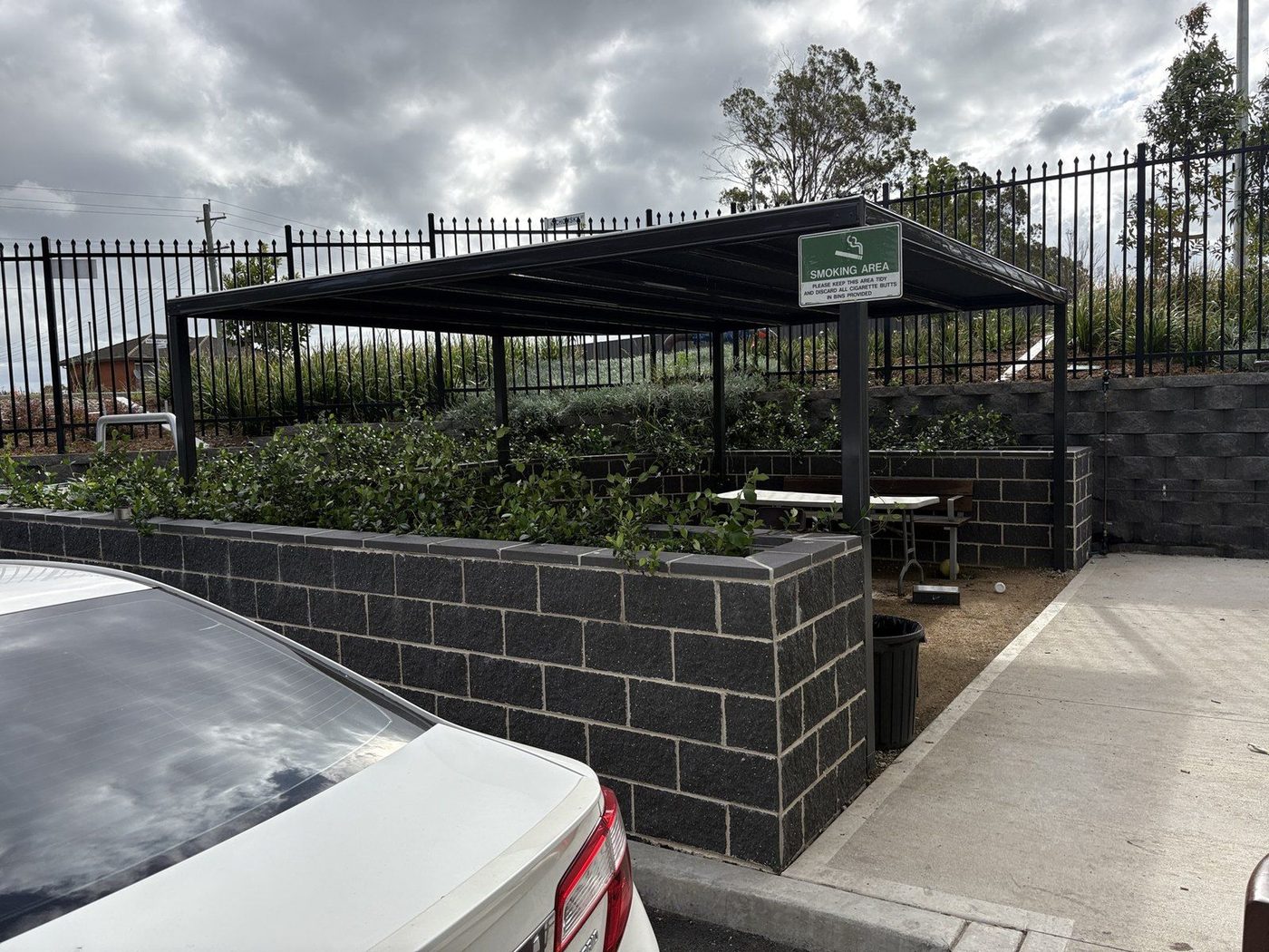 Flat black roof on steel posts over bench seating beside a block wall and car park., Leppington