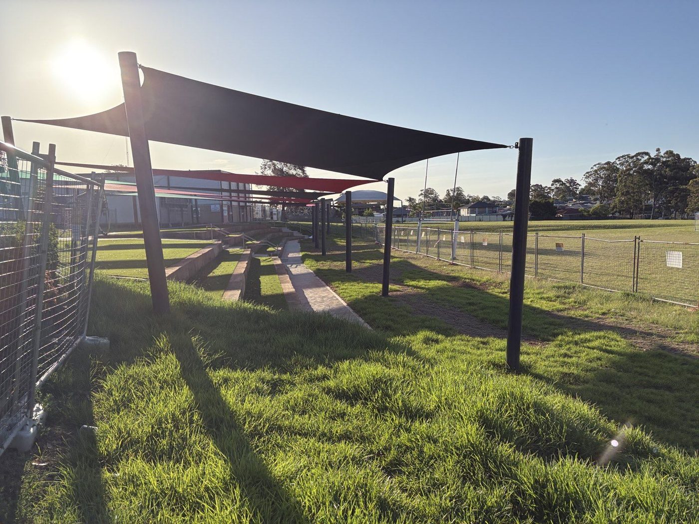 Black sail on dark posts shading concrete terrace seating beside a green sports field at sunset., Lurnea