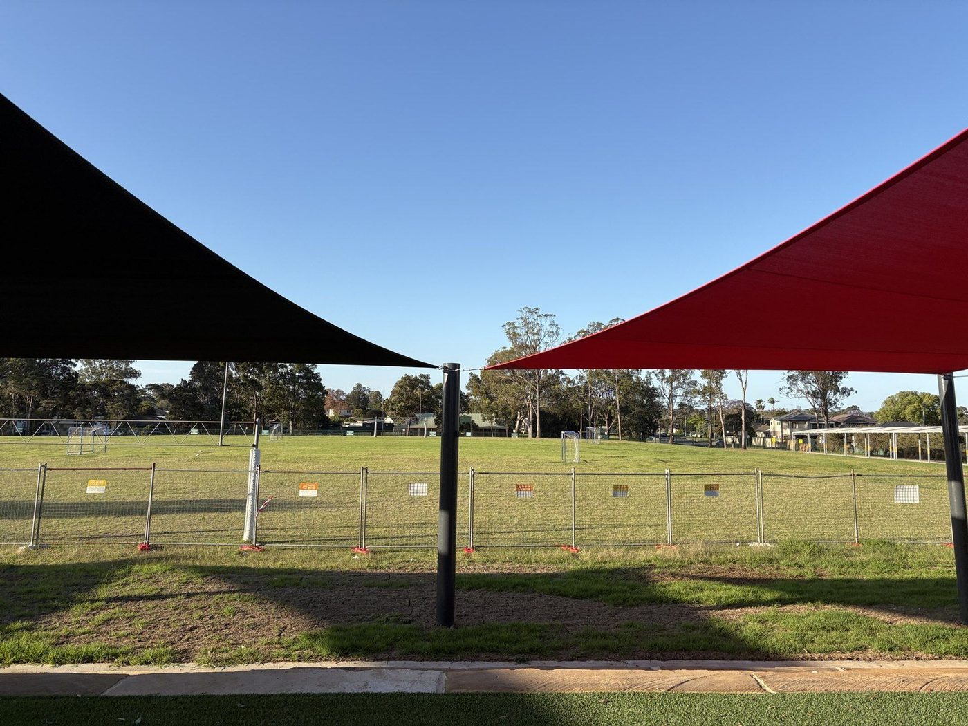 Black and red shade sails meeting at a steel post above a fenced grass sports field., Lurnea