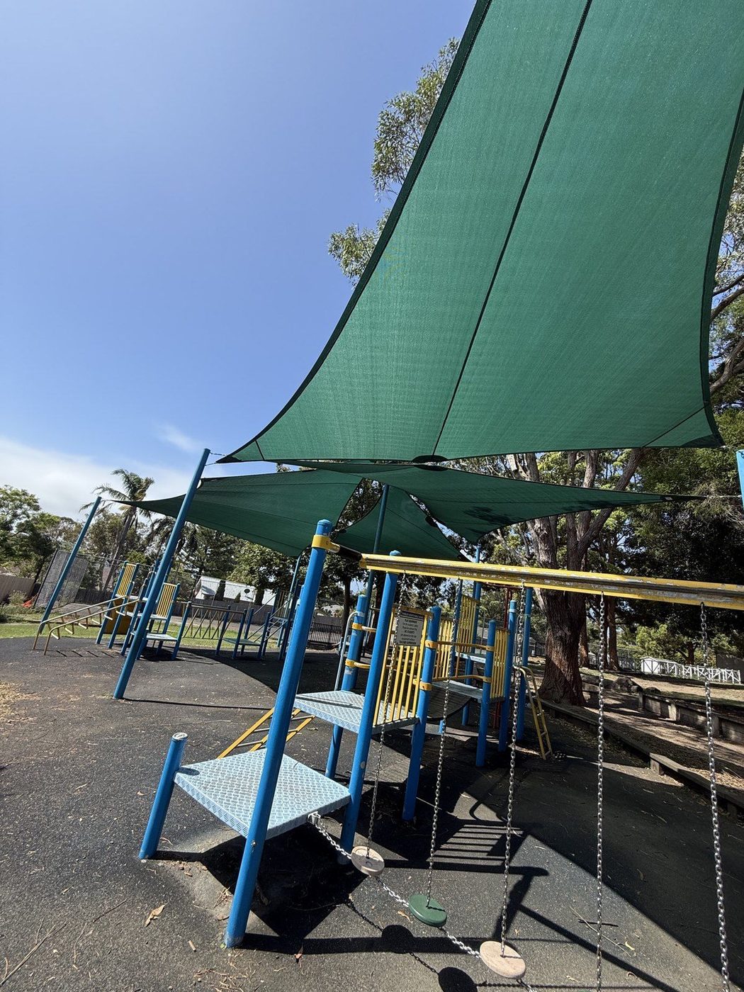 Green shade sails over playground equipment and swings, seen from below against a clear sky., Macquarie Park