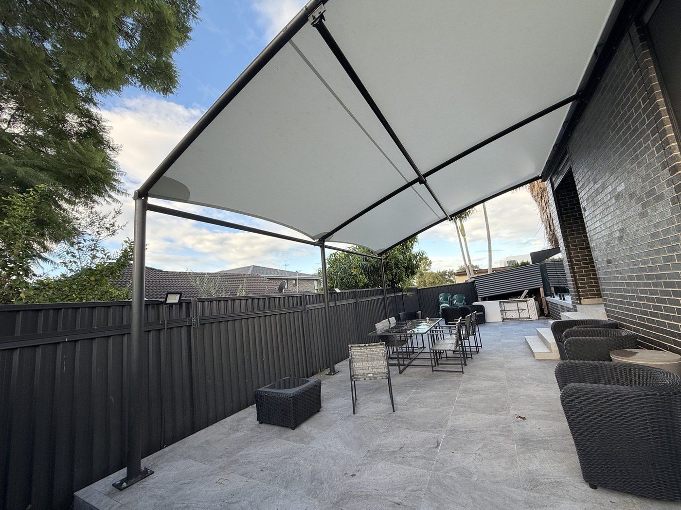 A white fabric canopy on a dark steel frame shading a tiled backyard patio with lounge seating., Merrylands West